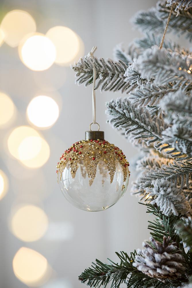 Clear glass Christmas ornament with beaded gold and red accents hanging on flocked pine branch