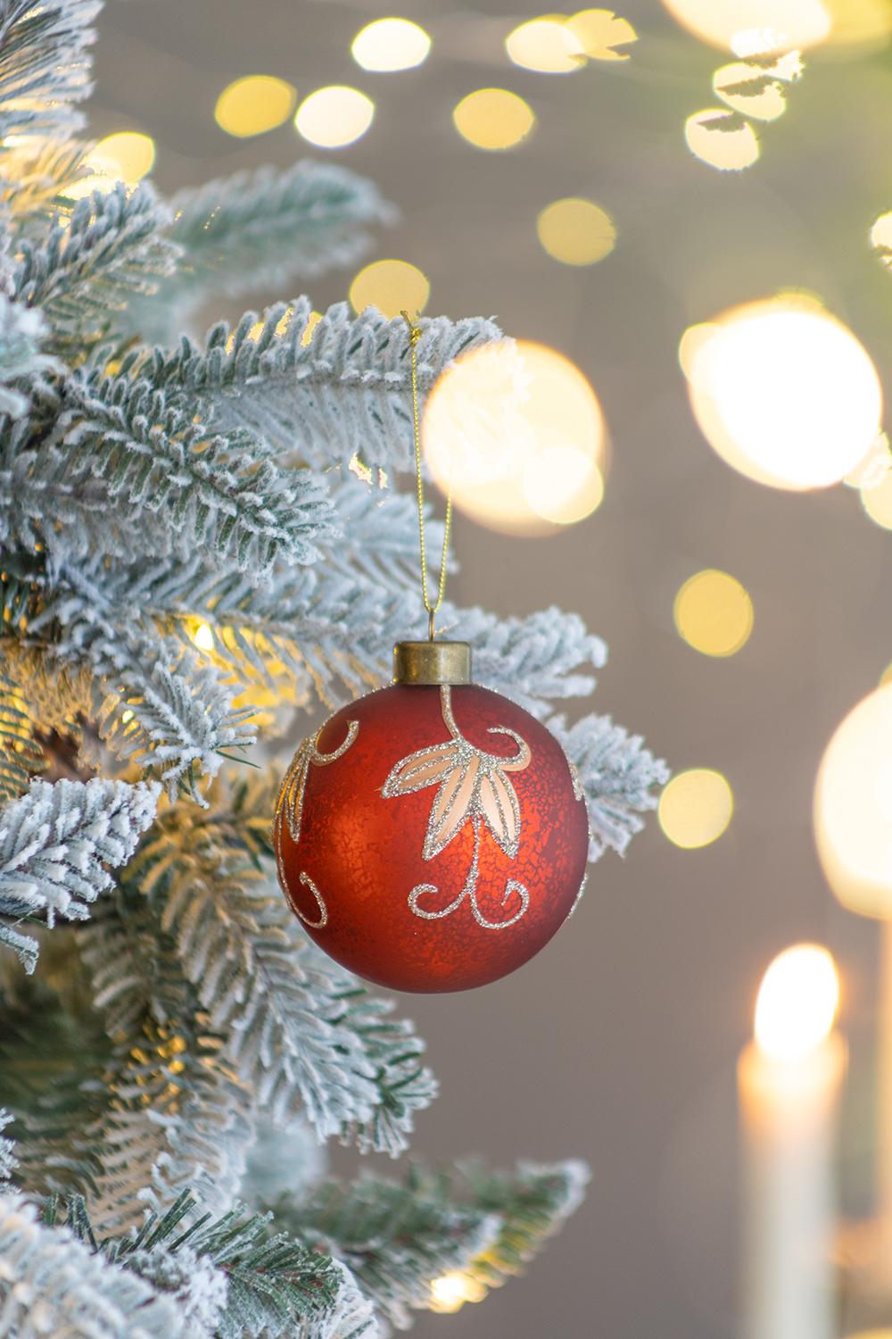 Glittering red glass Christmas ornament with gold vine detail hanging on frosted tree