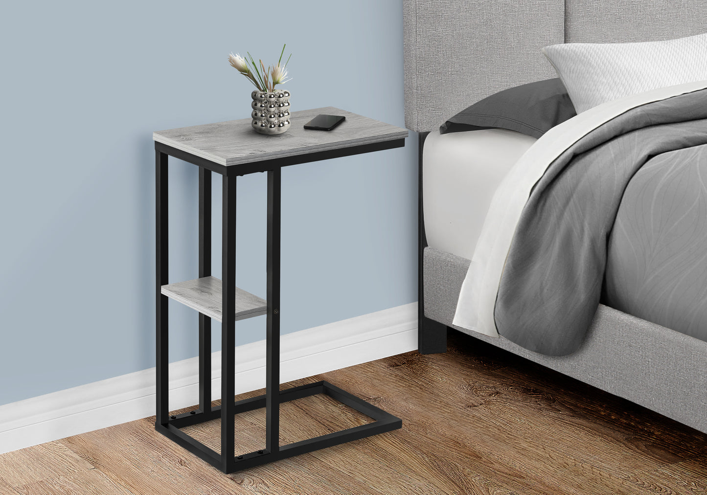 Cshaped end table with gray wood grain top and black metal frame next to a bed