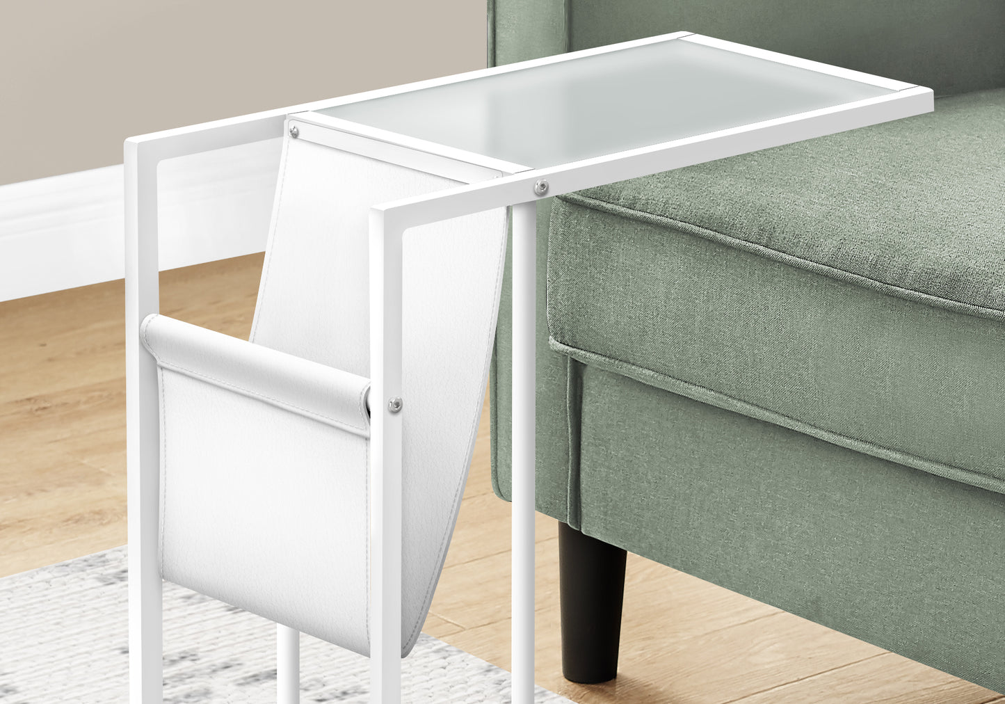 White metal C table with glass top and magazine holder beside green sofa