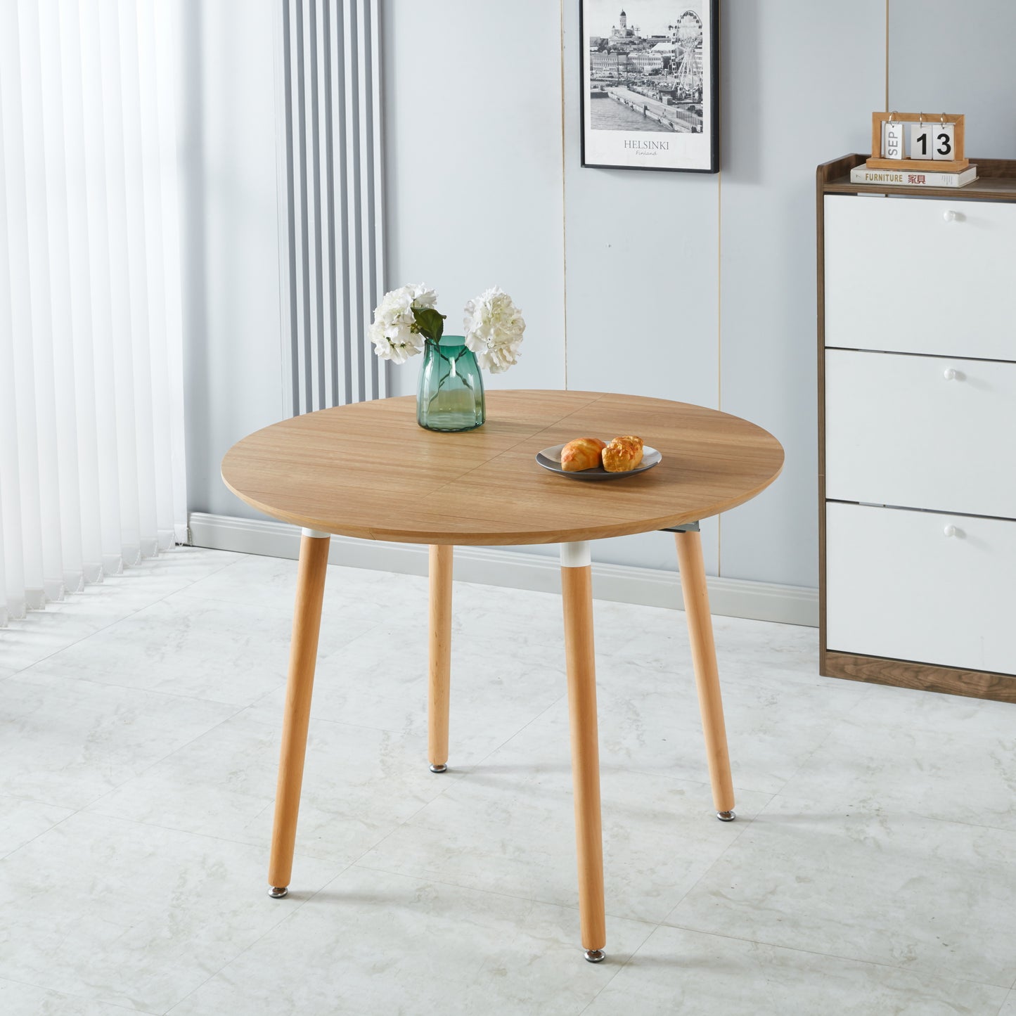 Round oak wood dining table with light wood legs featuring a vase of white flowers and pastries