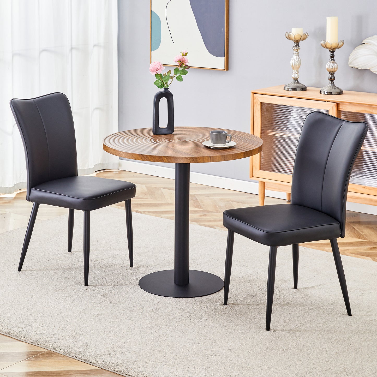Two black faux leather dining chairs with sleek metal legs flanking a round wood cafe table