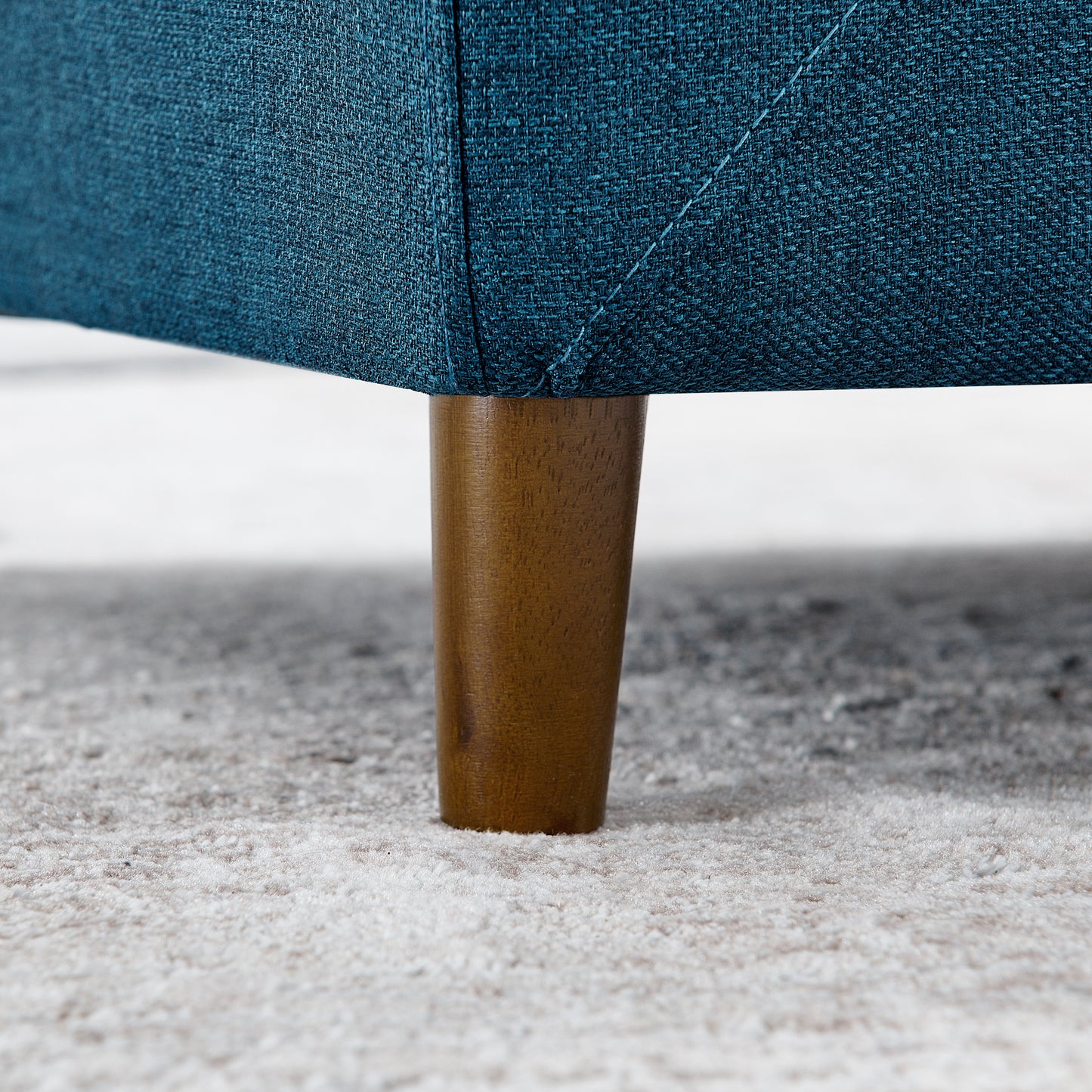 Closeup of a teal textured fabric sofa leg with a tapered wooden base on a neutral rug