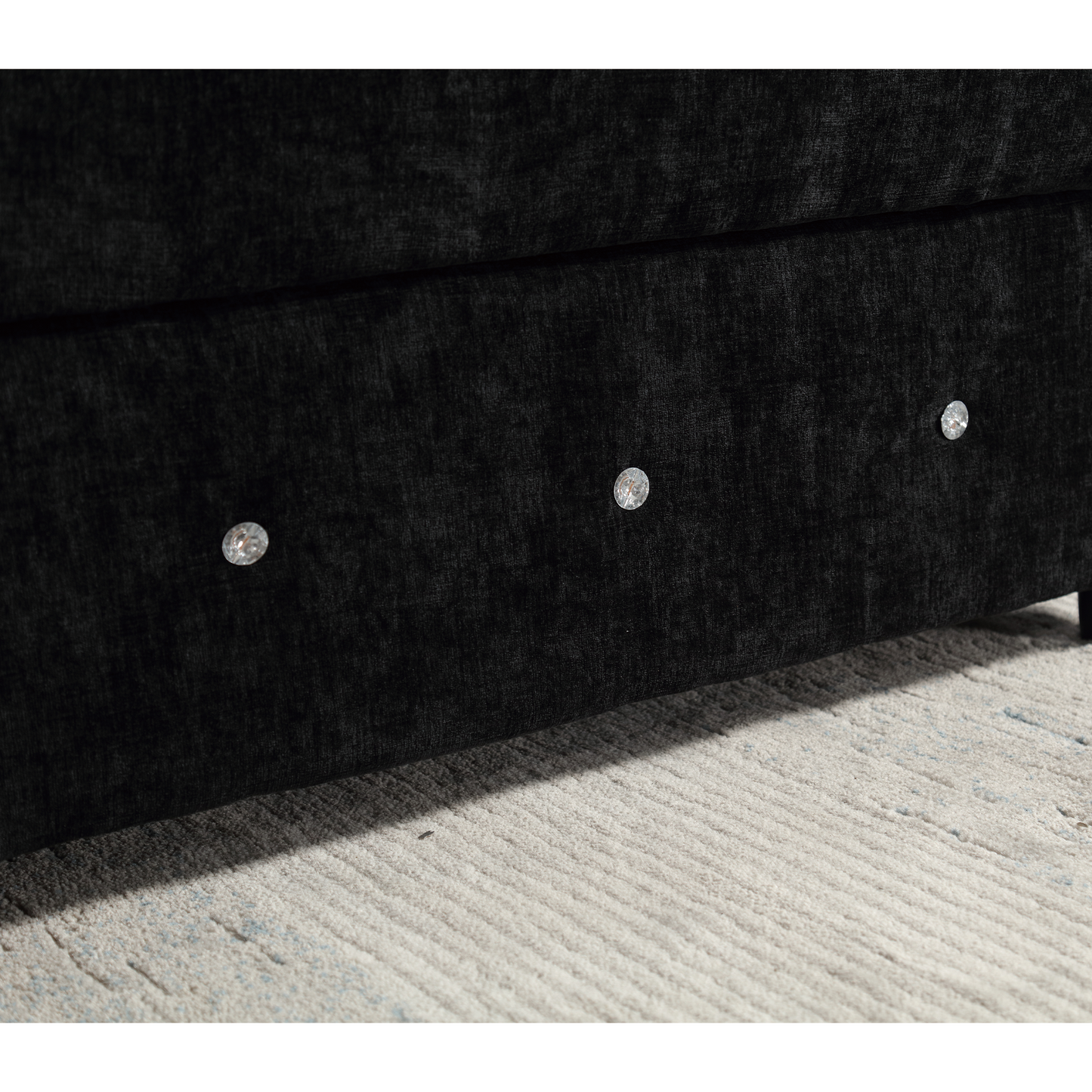 Closeup of a black velvet divan bed base with crystal button tufting on a light rug
