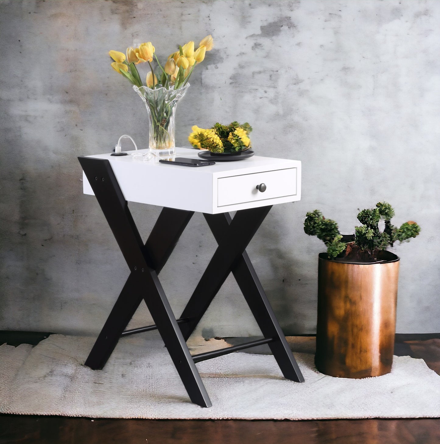 Modern white end table with black Xshaped legs and one drawer holding yellow tulips