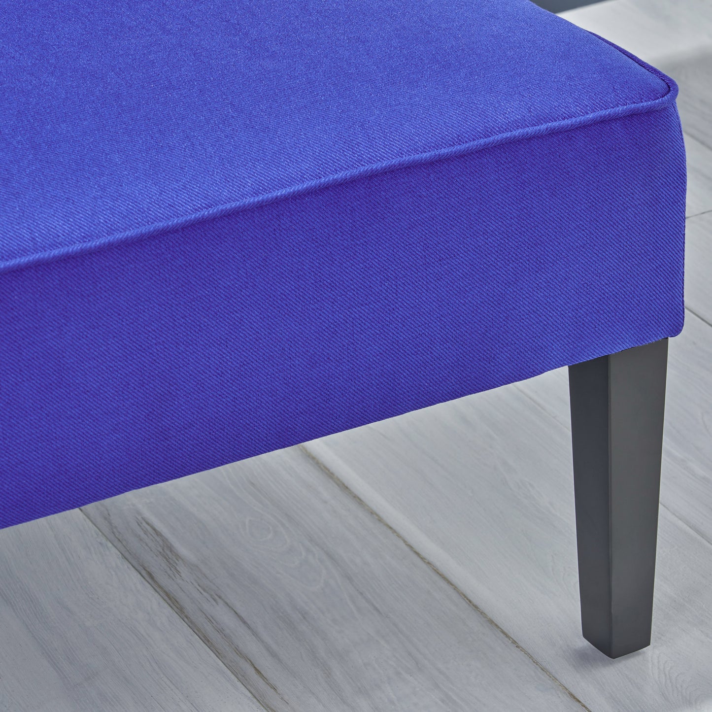 Close up of a rich royal blue upholstered ottoman with piped edges and dark tapered legs