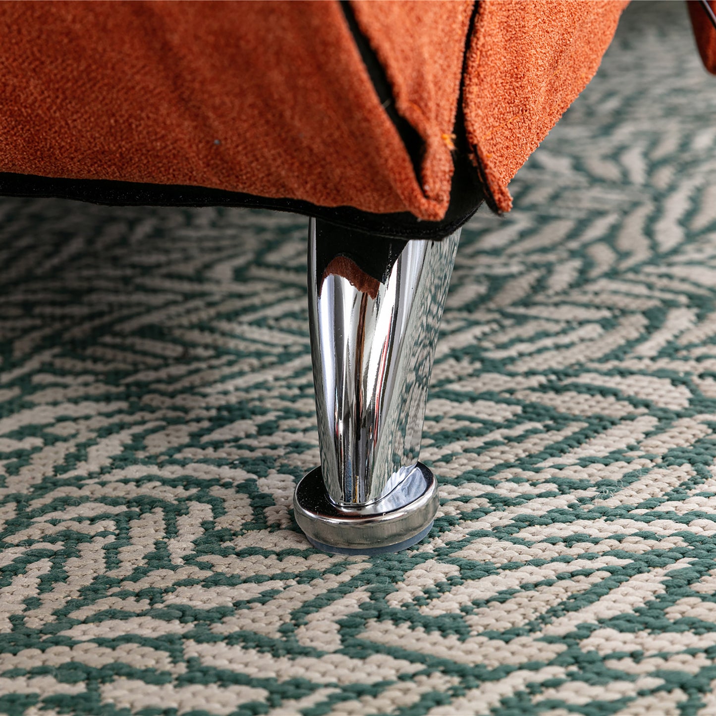 Orange suede sofa leg with a polished chrome finish resting on a geometric patterned rug