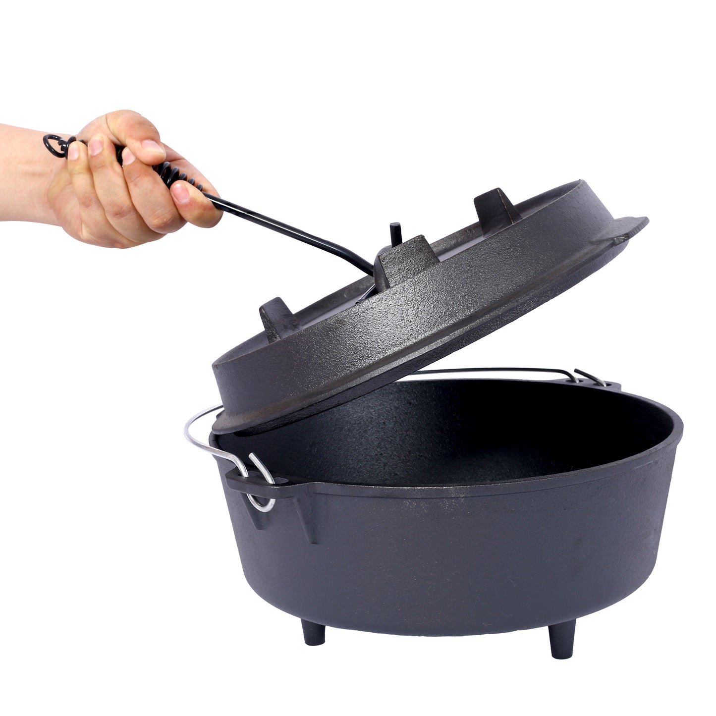 A hand lifts the lid of a black cast iron Dutch oven with legs ready for cooking