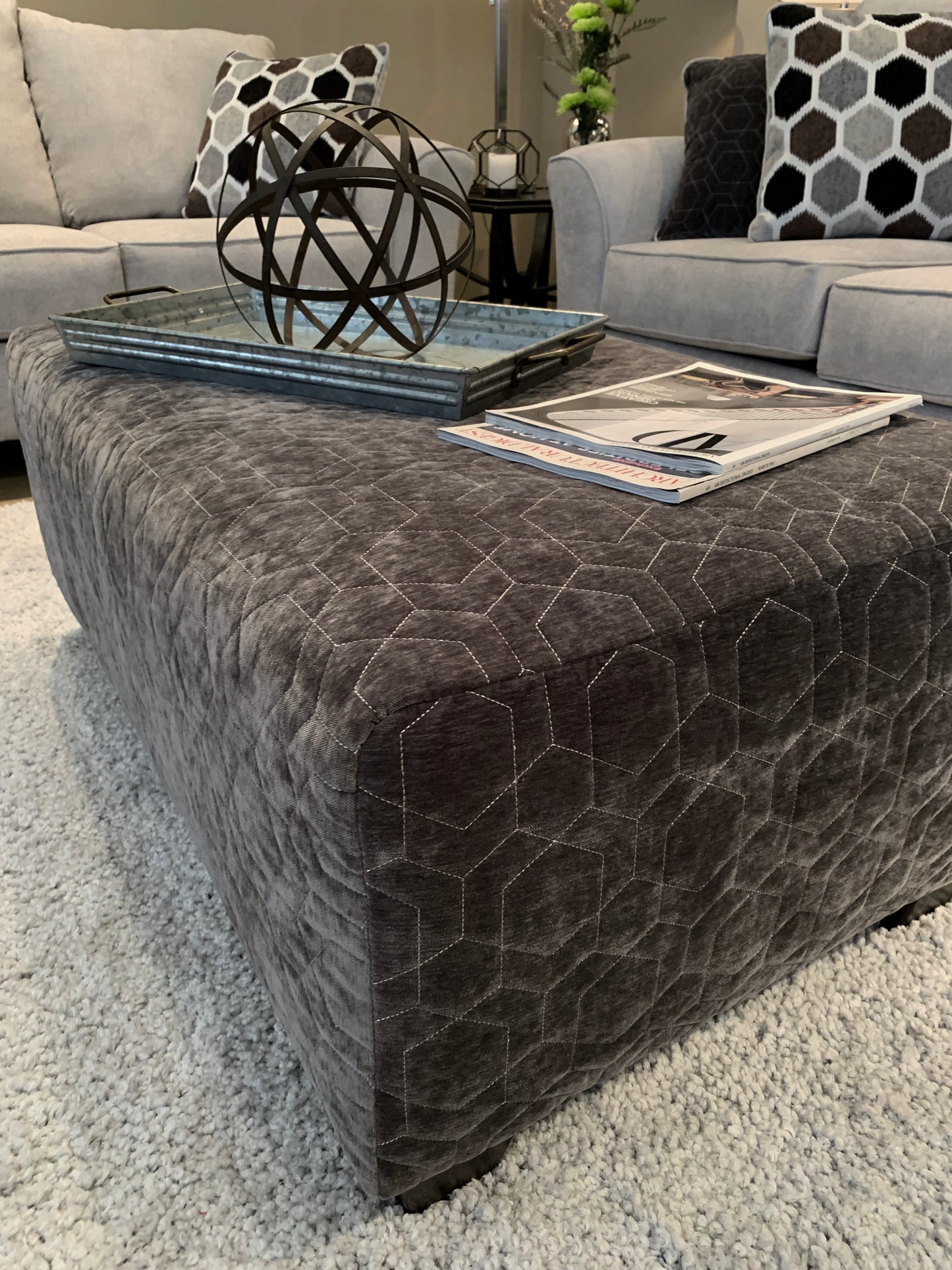 Dark gray velvet ottoman with hexagon quilting placed on a plush rug