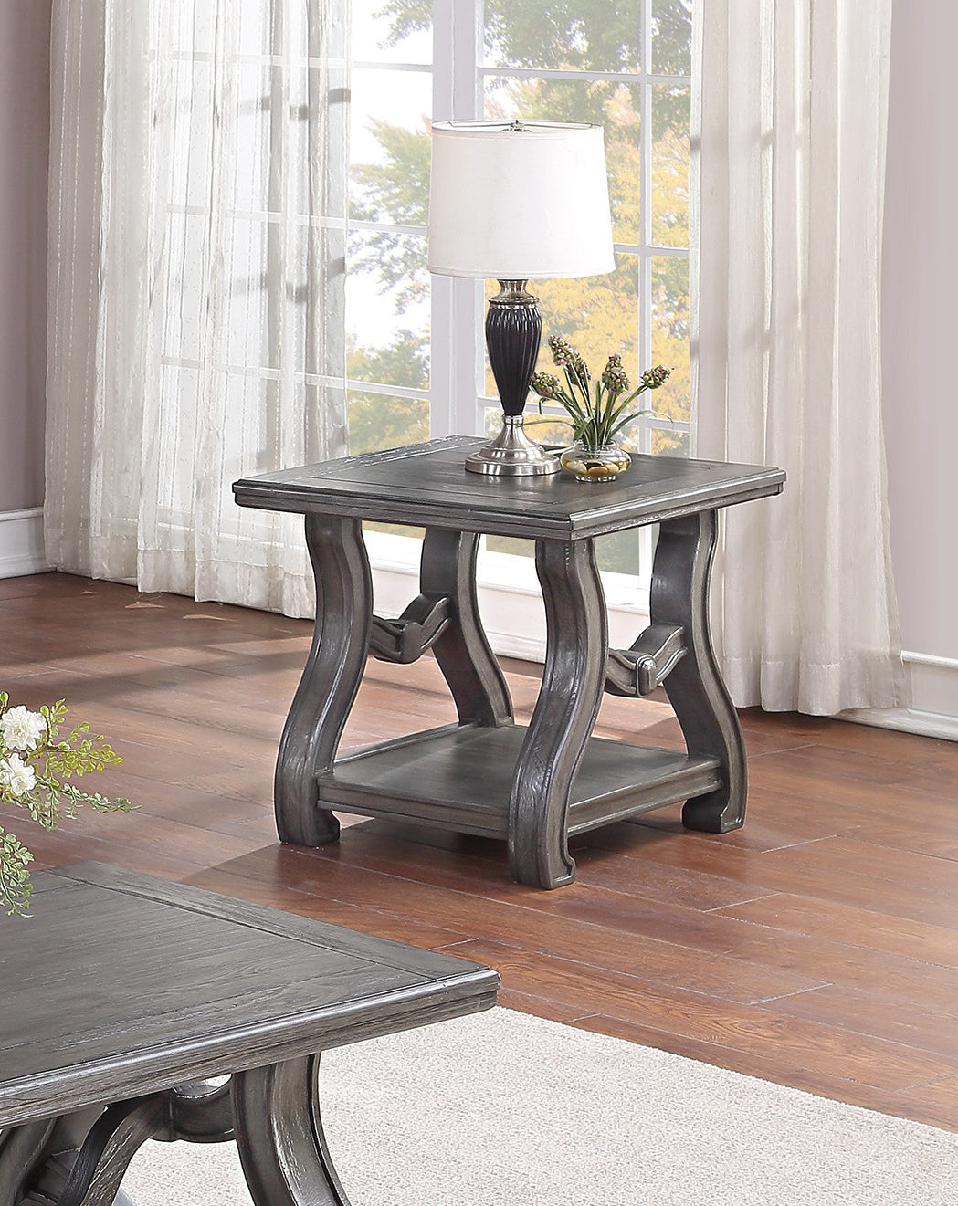 Gray wooden end table with carved legs and lower shelf holding a lamp and vase