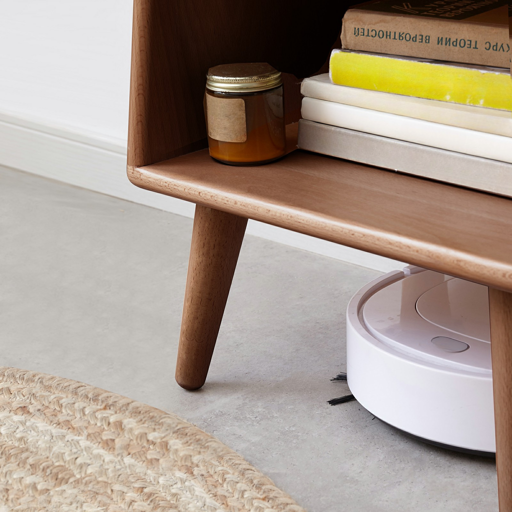 White circular robot vacuum cleaner with brushes cleaning under a wooden side table