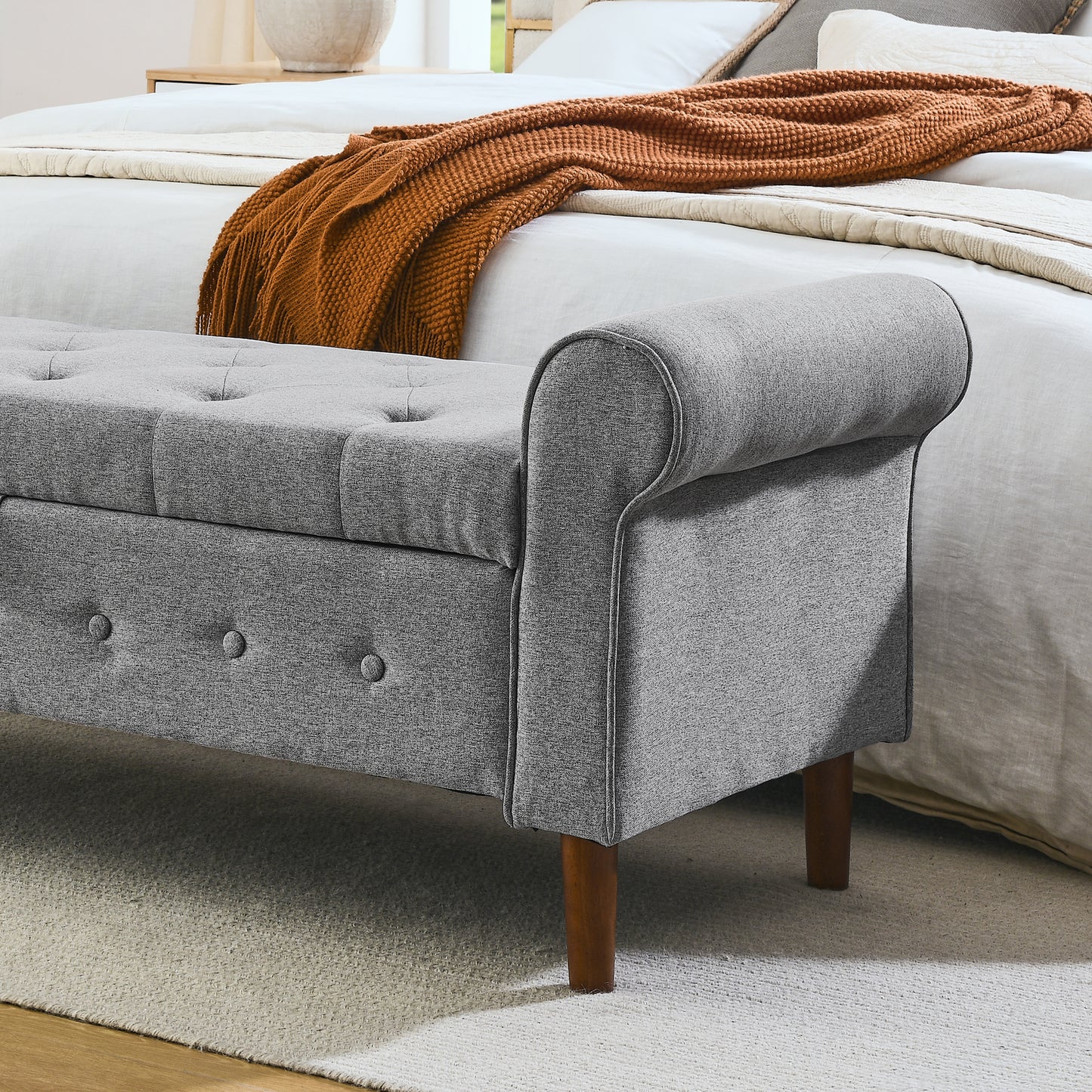 Grey tufted storage bench with rolled arms and dark wood legs at foot of bed