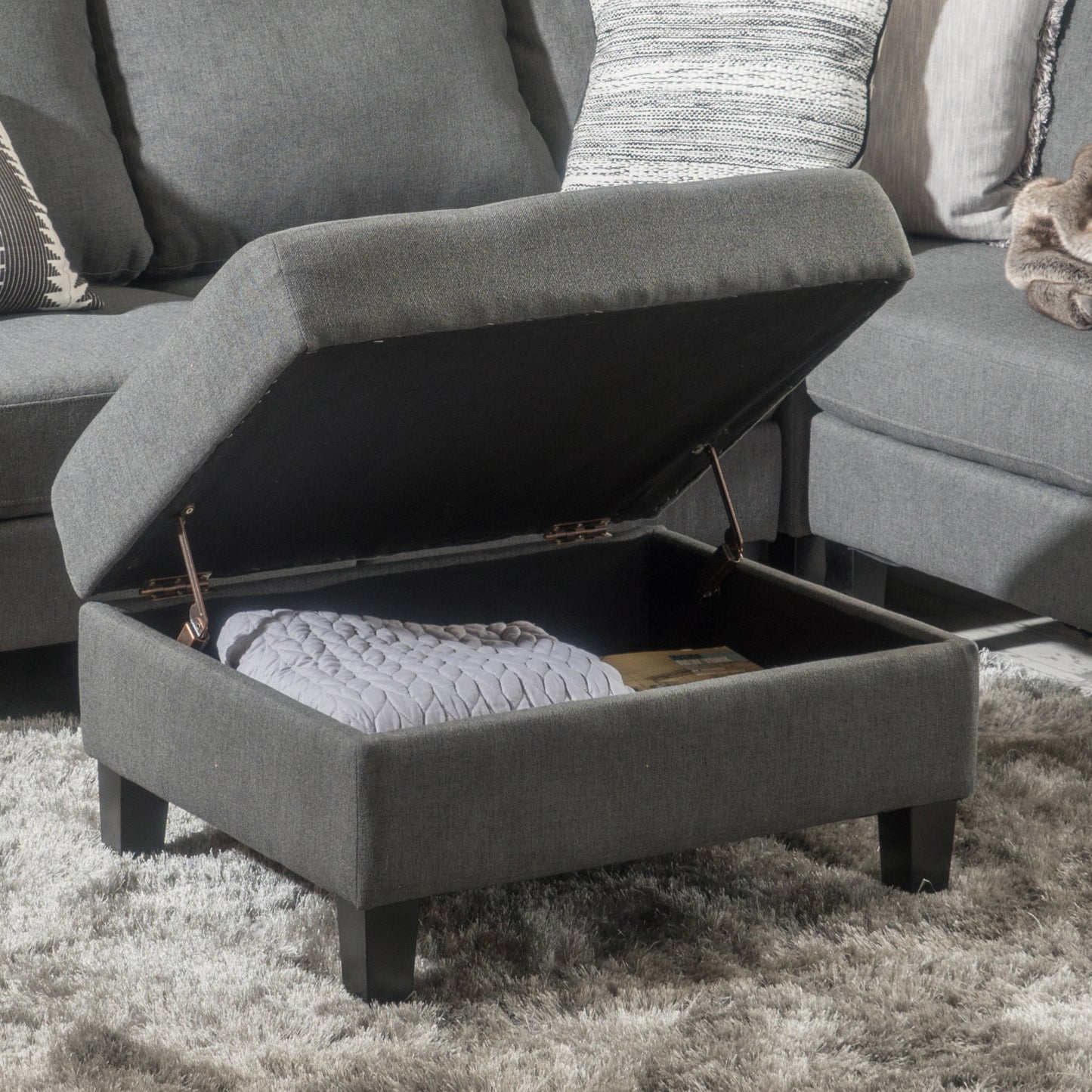 Dark grey fabric storage ottoman with lid open showing blanket and book inside