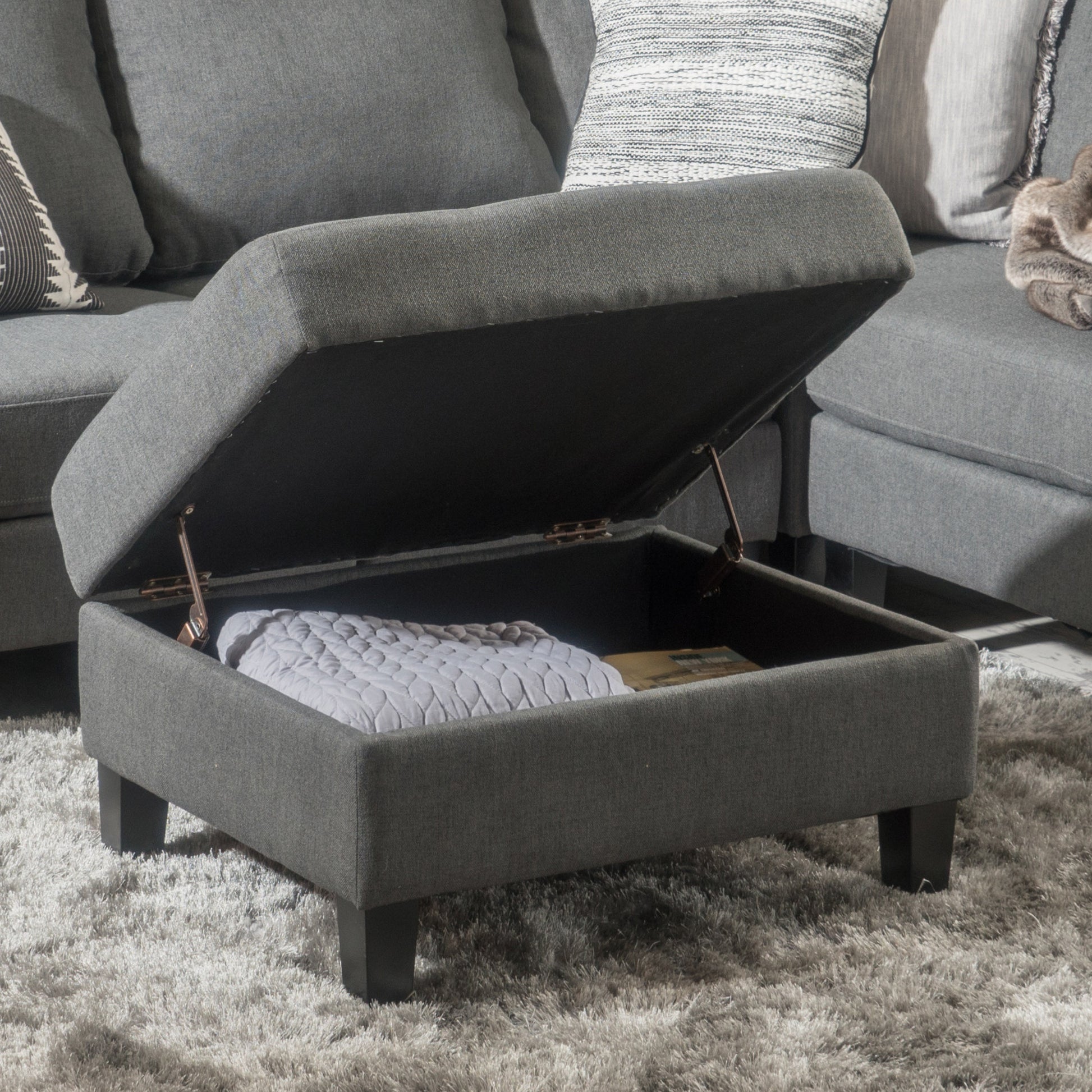 Dark grey fabric storage ottoman with lid open showing blanket and book inside