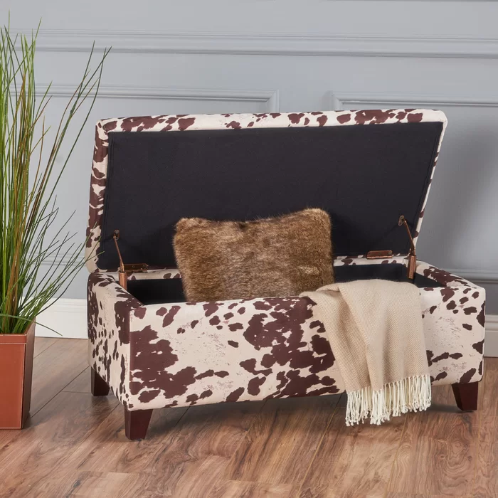 Cow print storage ottoman with dark brown legs and open lid showing dark interior and throw pillow