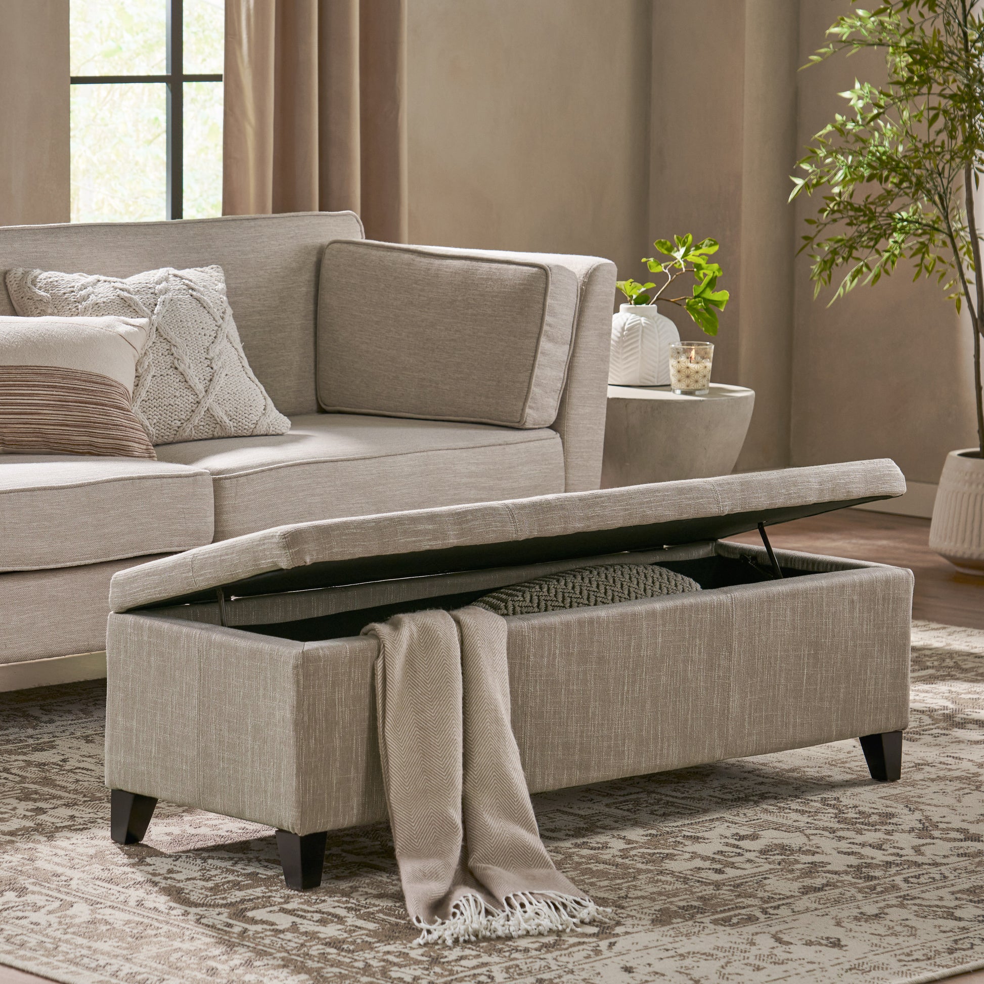 Beige upholstered storage ottoman with open lid revealing blanket storage