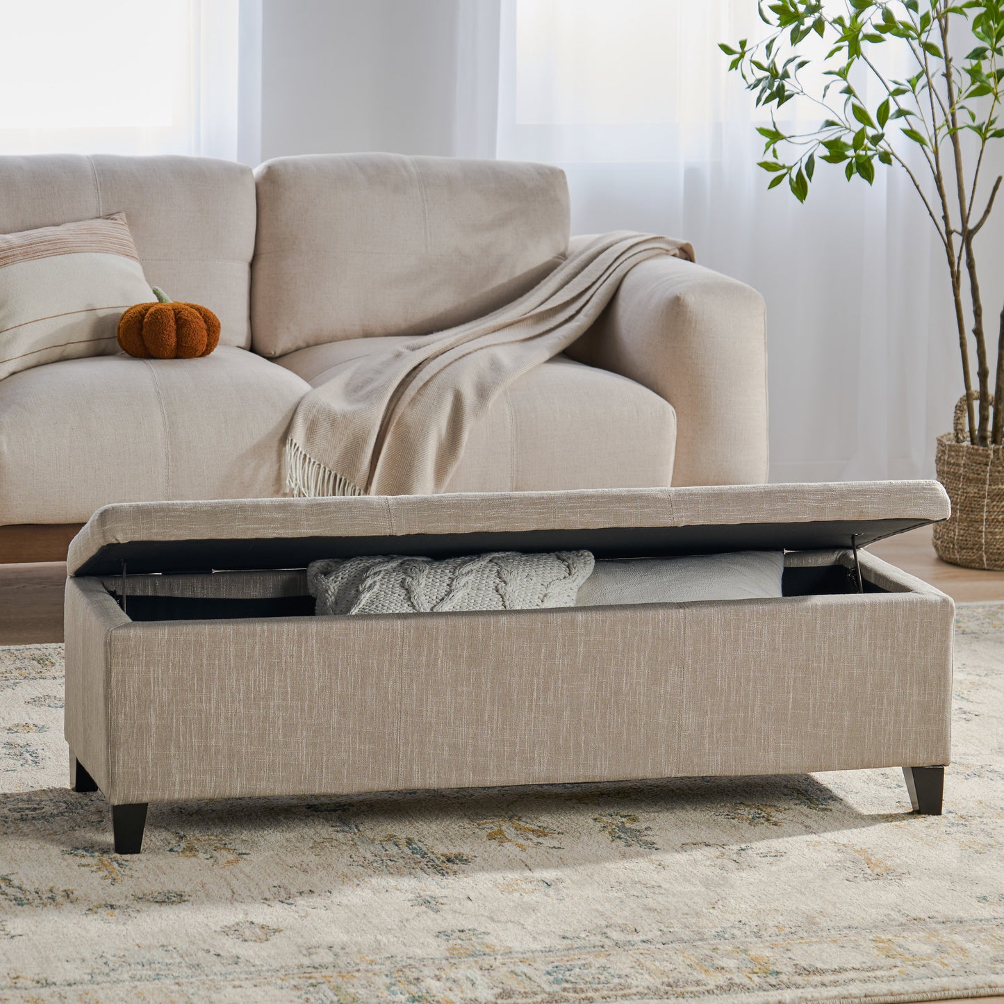 Beige linen storage ottoman with lifted lid revealing blankets