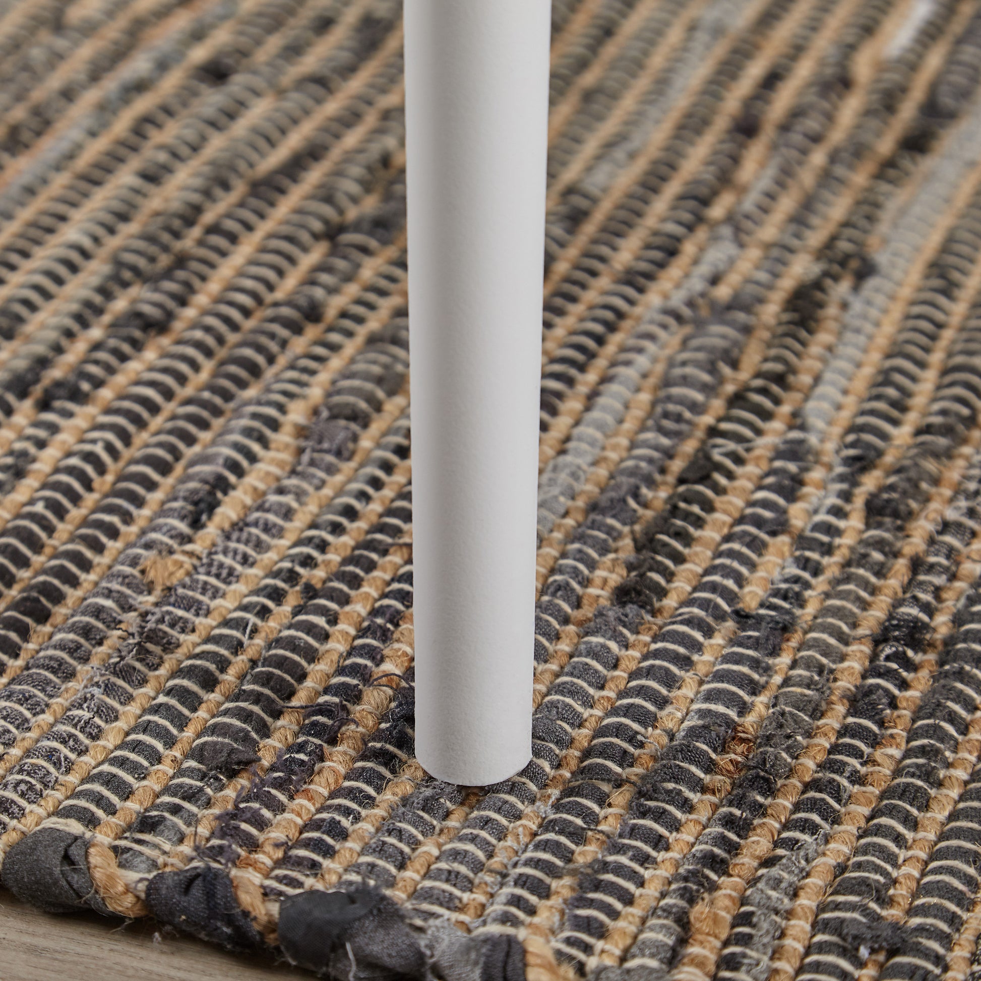 Closeup of a white cylindrical leg resting on a textured braided rug with grey and tan woven fibers