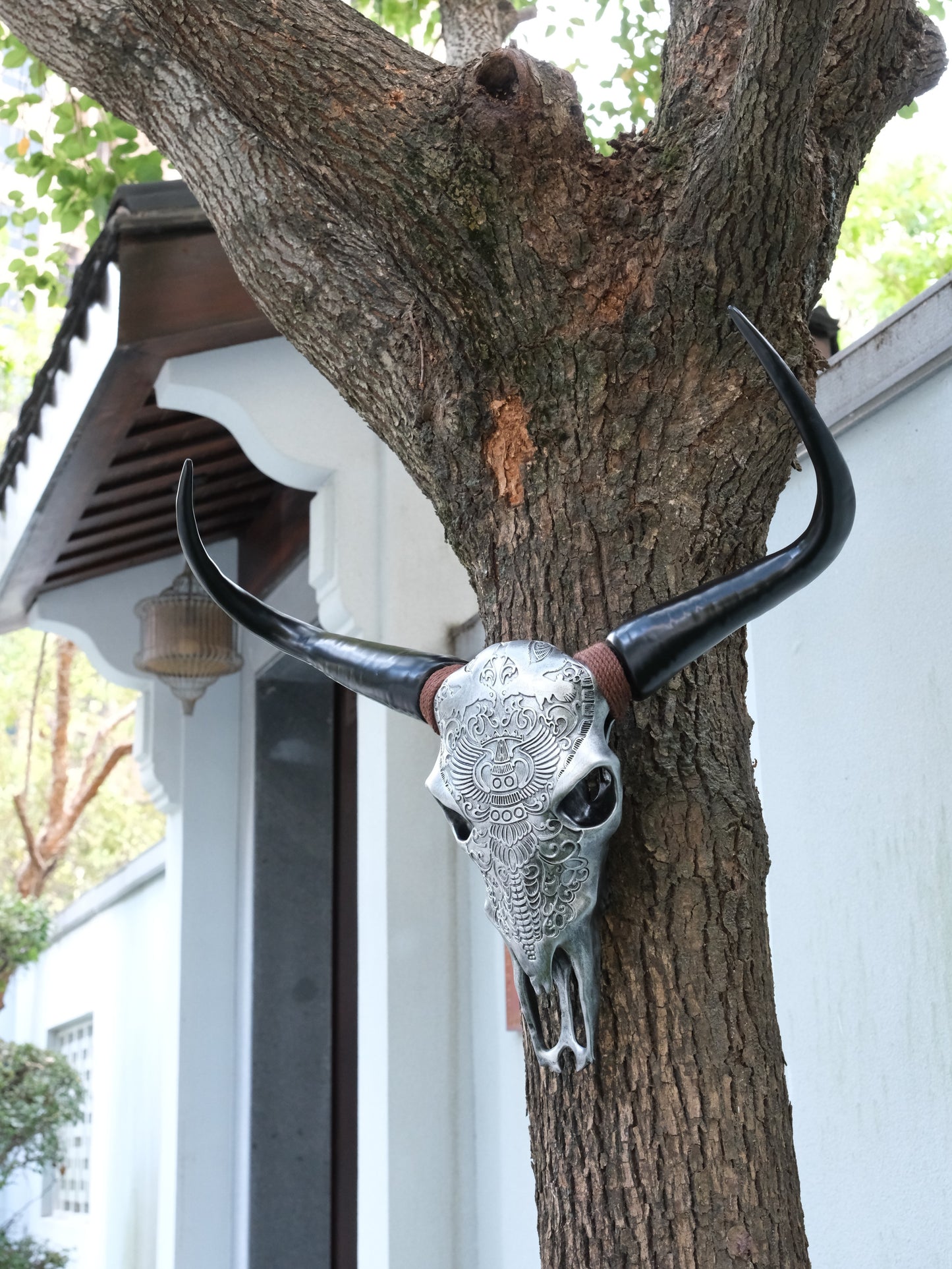 Ornate silver bull skull decoration with black horns mounted on a tree trunk for outdoor decor