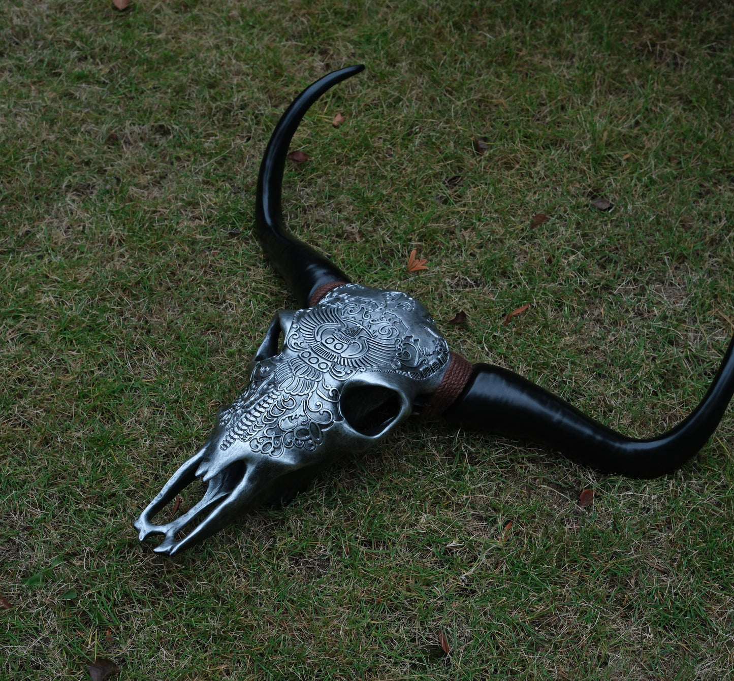 Silver bull skull replica with ornate relief carvings and black horns on grass