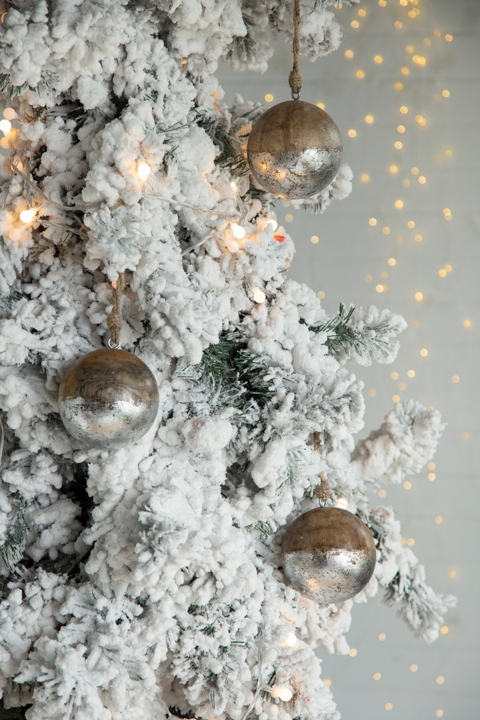 Flocked Christmas tree branches with three silver and wood ornamental baubles and warm LED string lights
