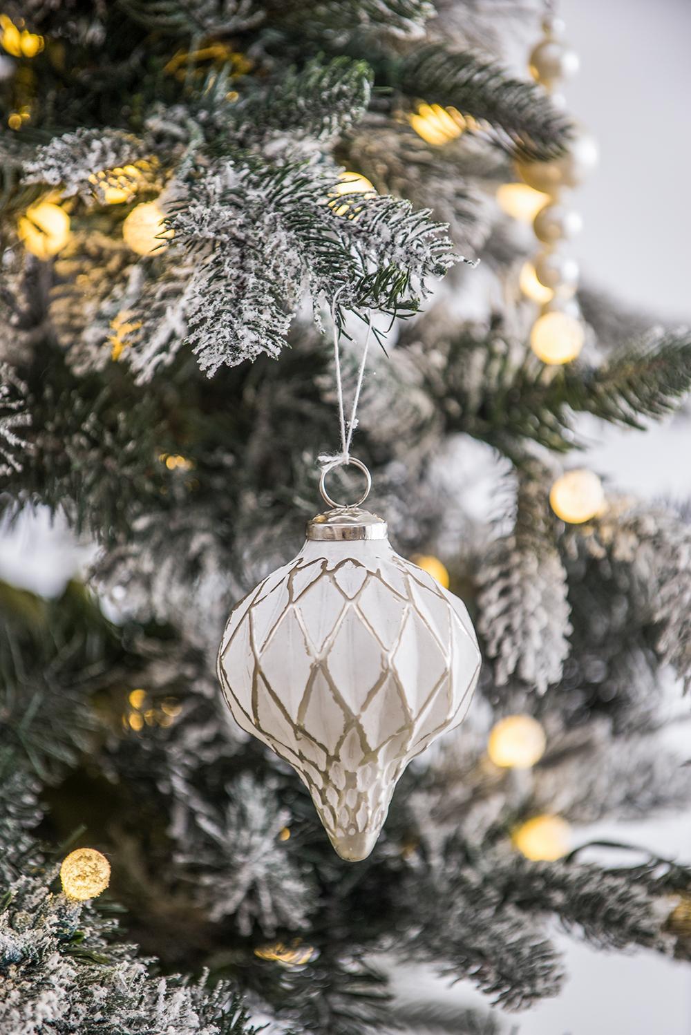 White ornate glass Christmas ornament with diamond pattern hanging from frosted tree branch