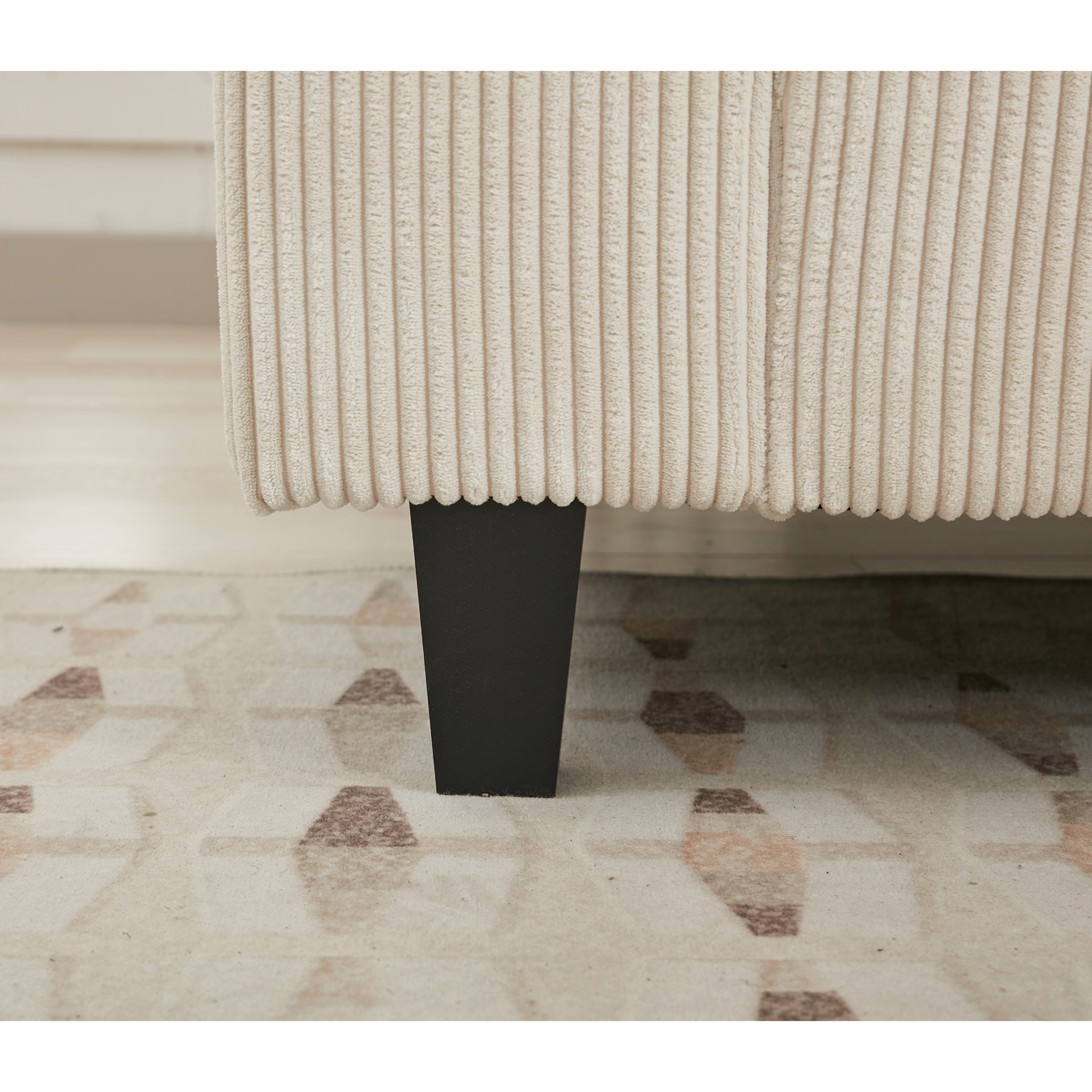 Closeup of a beige corduroy sofa leg with a black tapered foot on a patterned rug