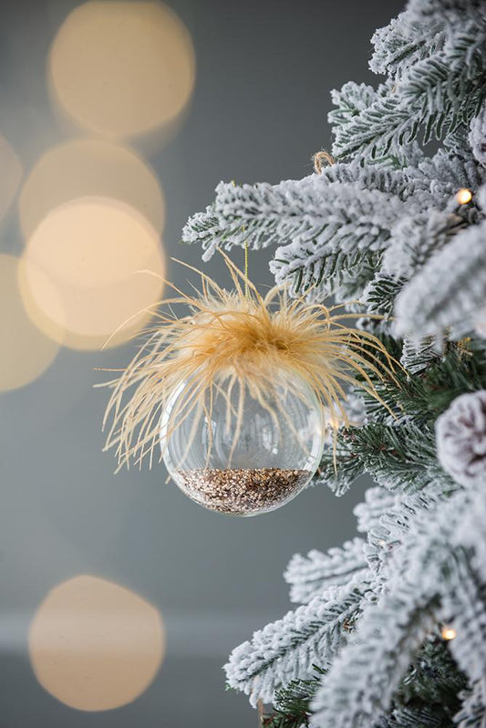 Clear glass bauble with tan feathers and glitter filling on a snowy Christmas tree branch
