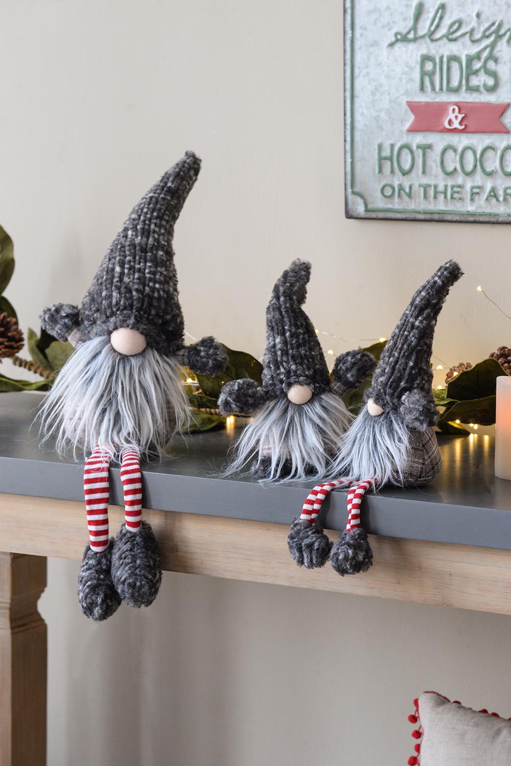 Three plush Christmas gnomes with grey knitted hats and fluffy white beards sit on a shelf