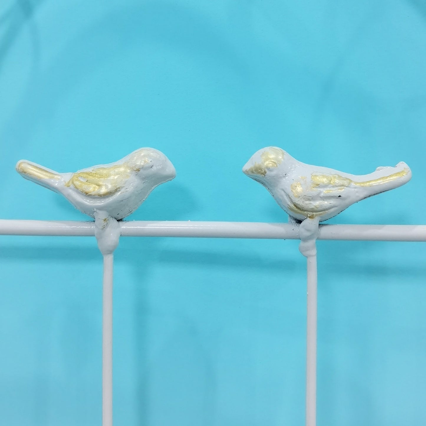 Two light blue and gold decorative bird figurines perched on a white railing against a teal background