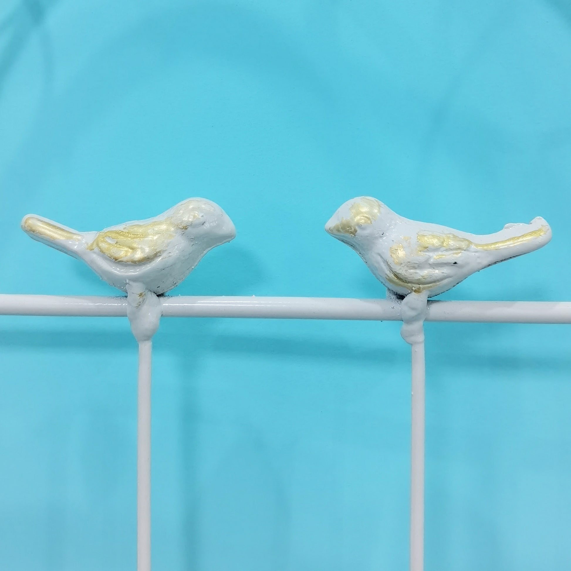 Two light blue and gold decorative bird figurines perched on a white railing against a teal background