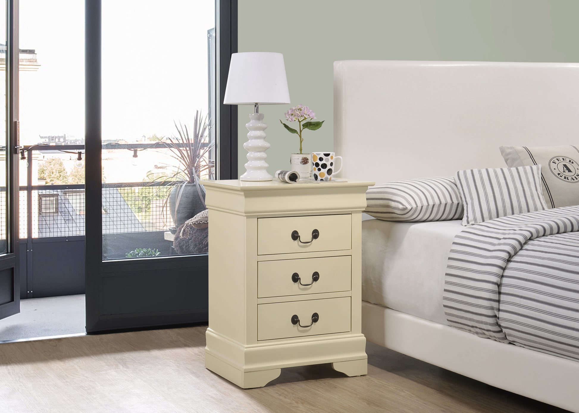 Cream wooden bedside table with three drawers and brass handles next to a white bed