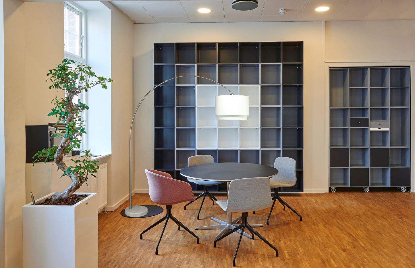 Black round meeting table with four modern swivel chairs a floor lamp and a large wall shelving unit