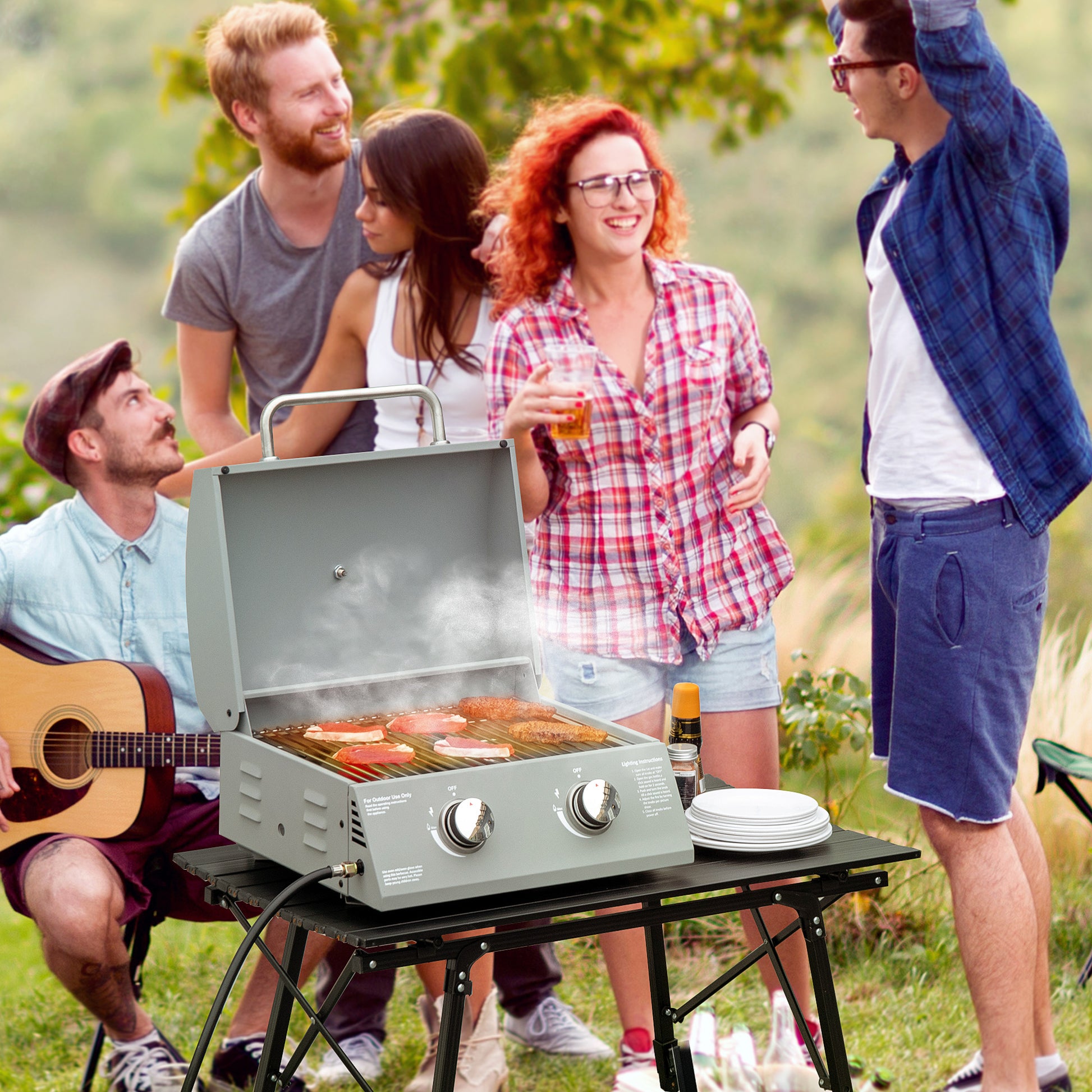 Grey twoburner portable propane grill cooking steaks outdoors with friends playing guitar