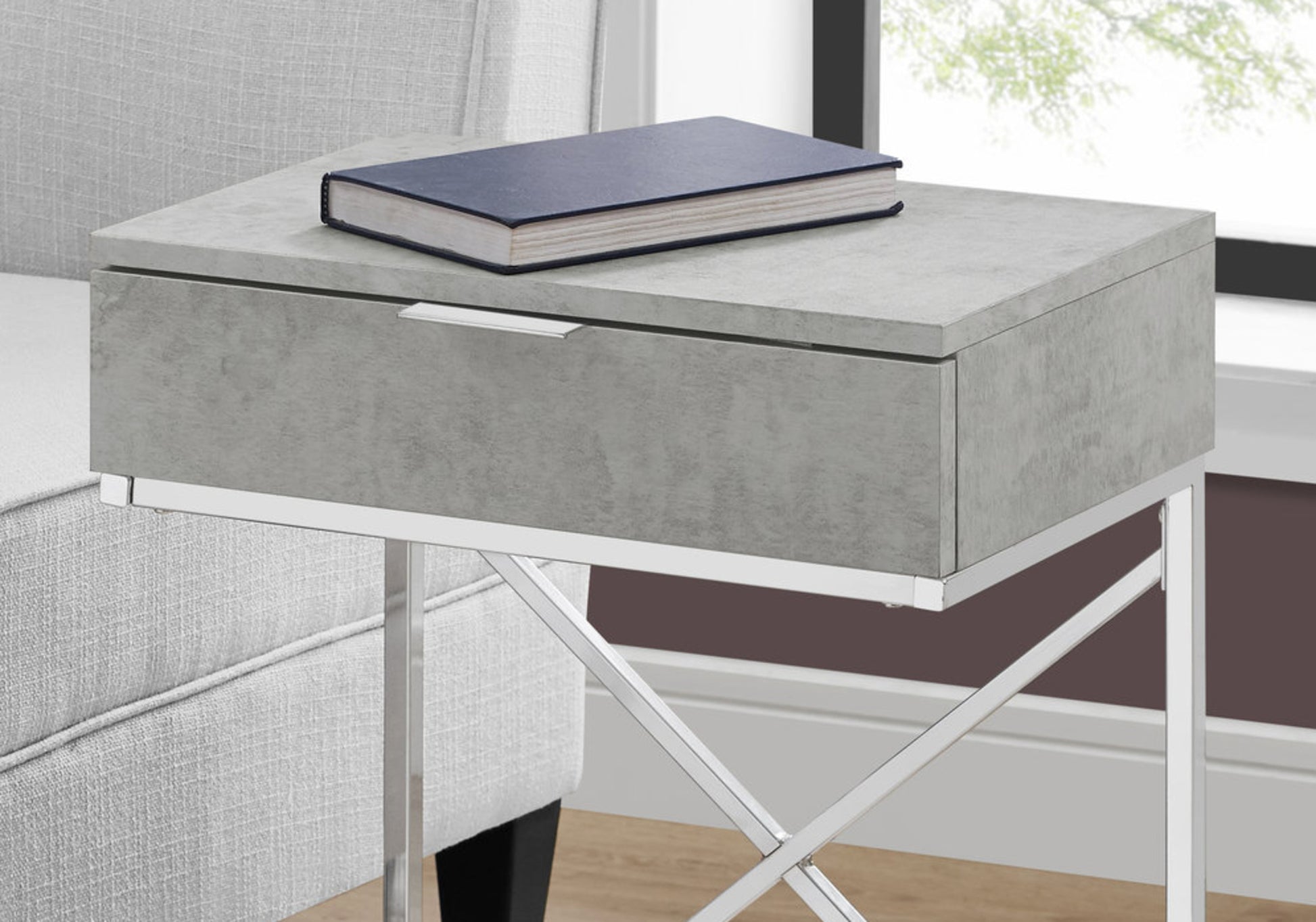 Modern concrete finish side table with single drawer and silver hardware next to armchair