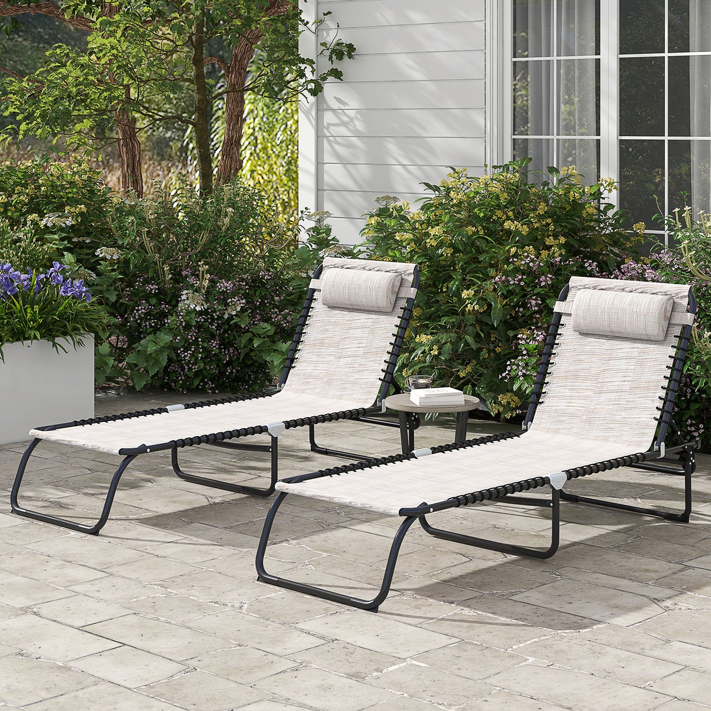Two beige outdoor chaise lounge chairs with black frames sit on a patio next to lush greenery