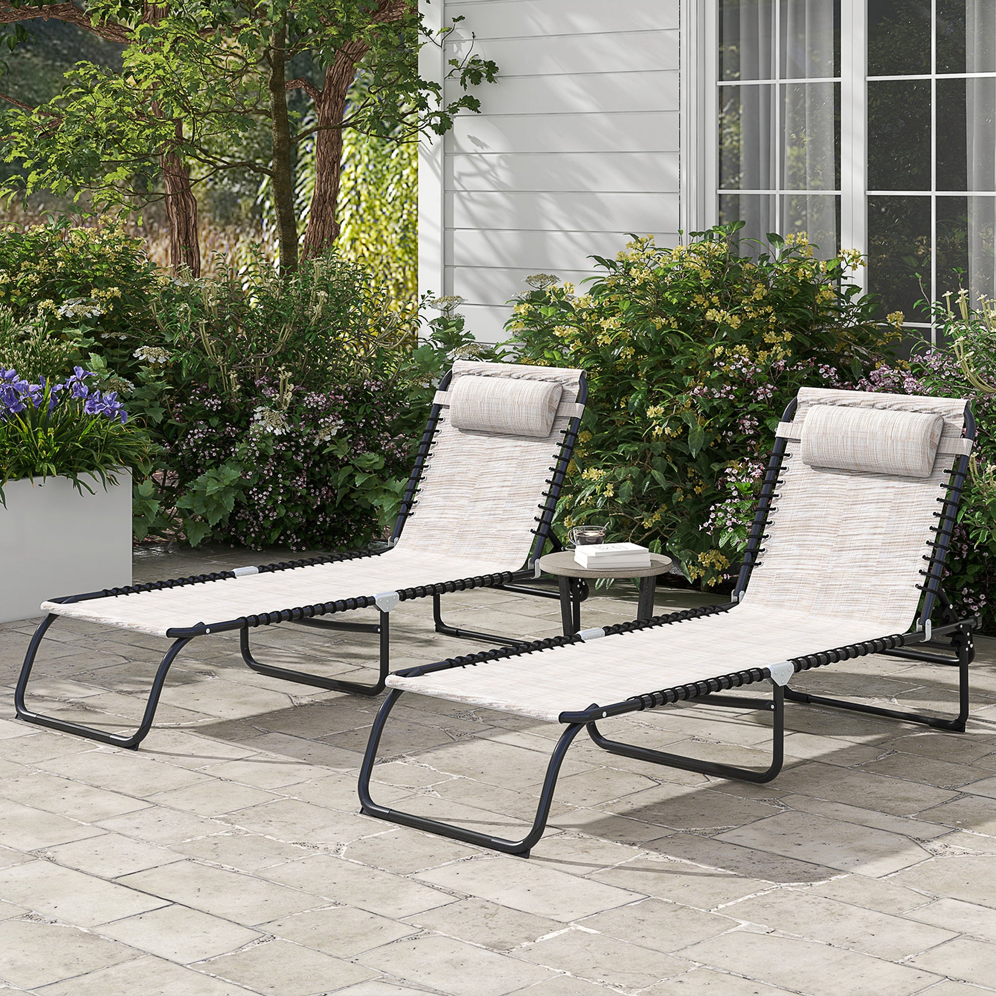 Two beige outdoor chaise lounge chairs with black frames sit on a patio next to lush greenery
