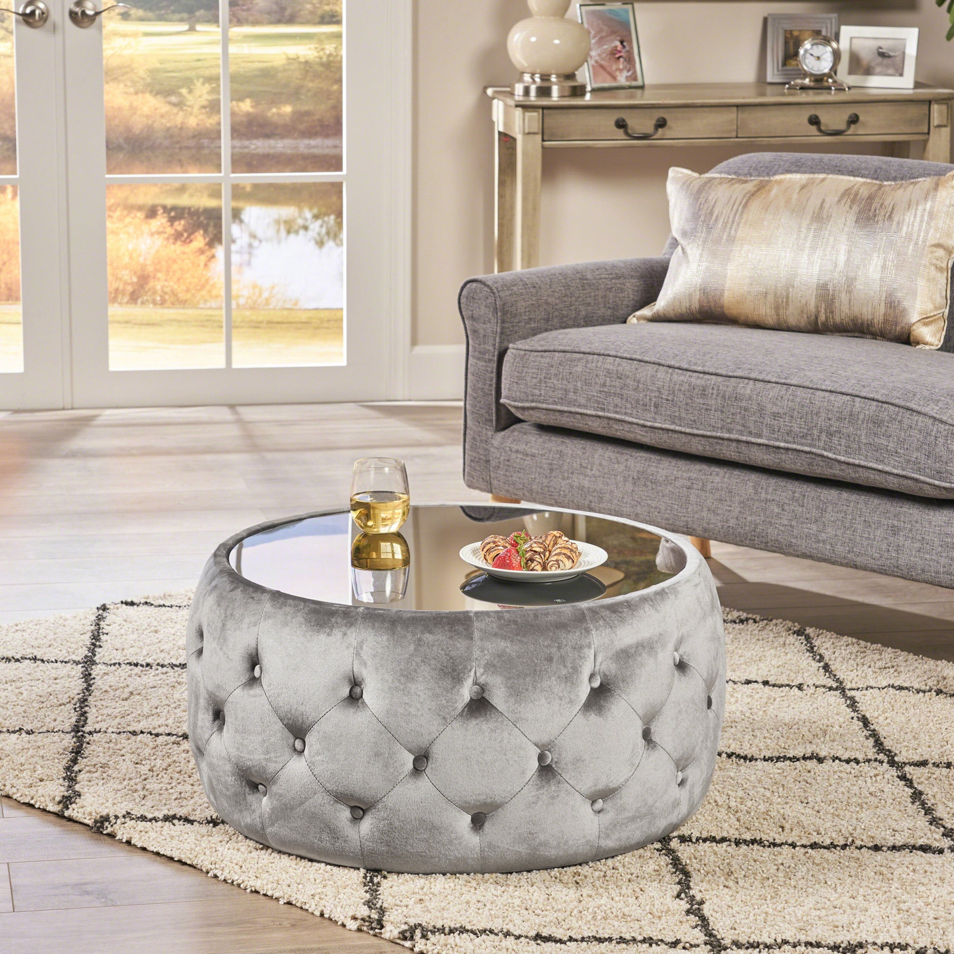 Tufted grey velvet ottoman coffee table with mirrored top and glass of drink