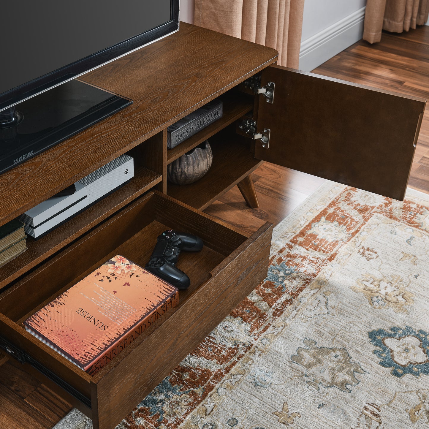 Dark oak entertainment center with open shelves and a drawer holding a black video game controller and a book titled Sunrise and Sunsets