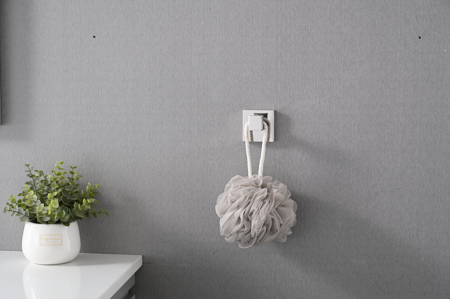 Soft beige mesh shower pouf with white rope handle hanging from a modern square hook