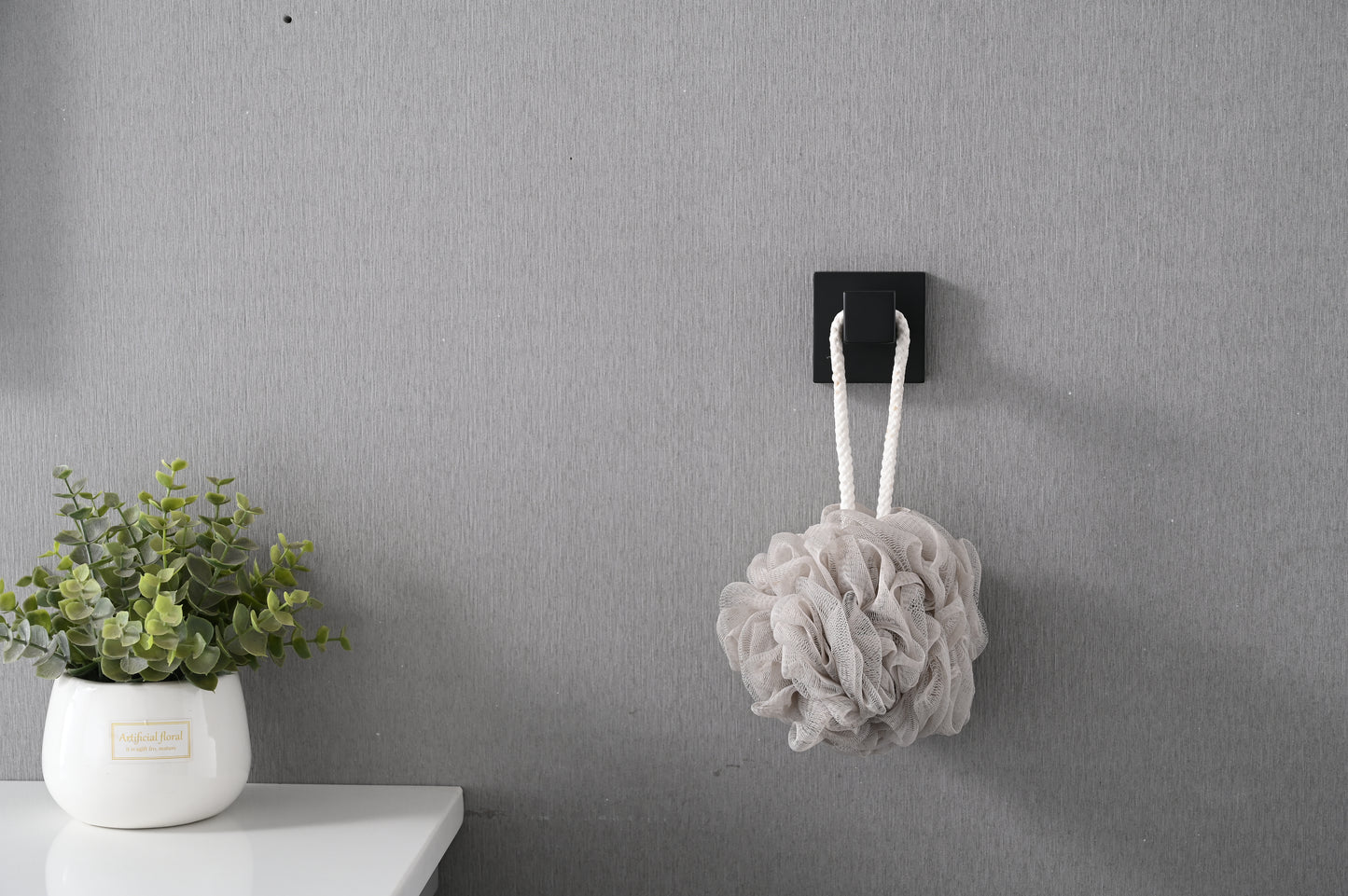 Plush grey bath loofah with white rope hanger against a textured wall