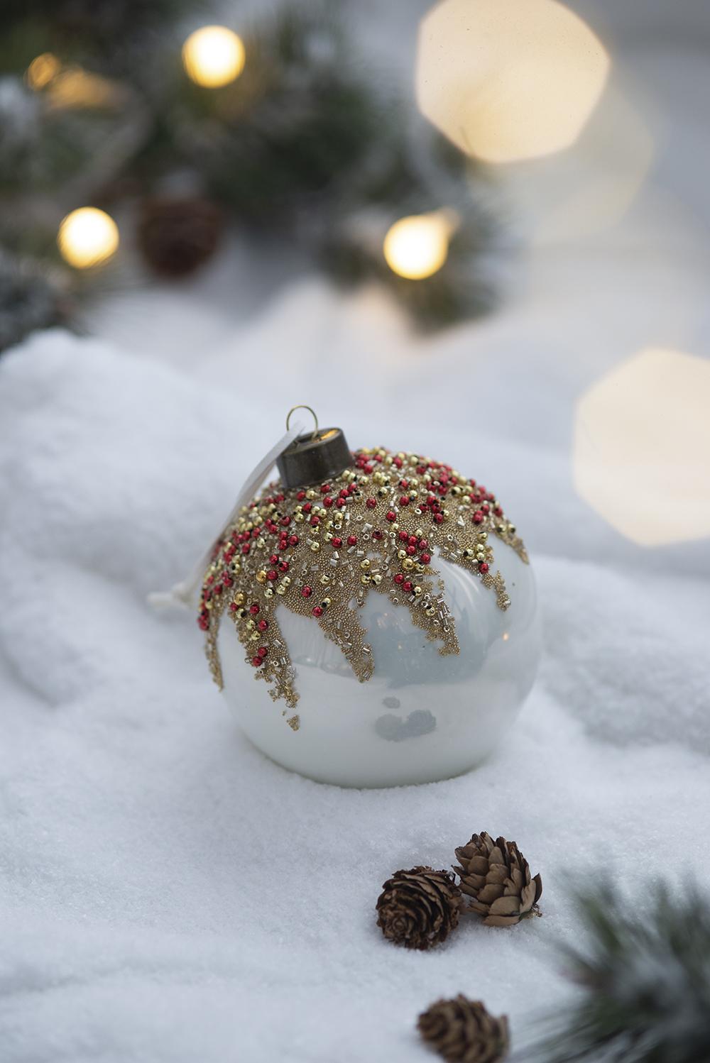 White Christmas ornament with gold and red beaded accents resting in artificial snow