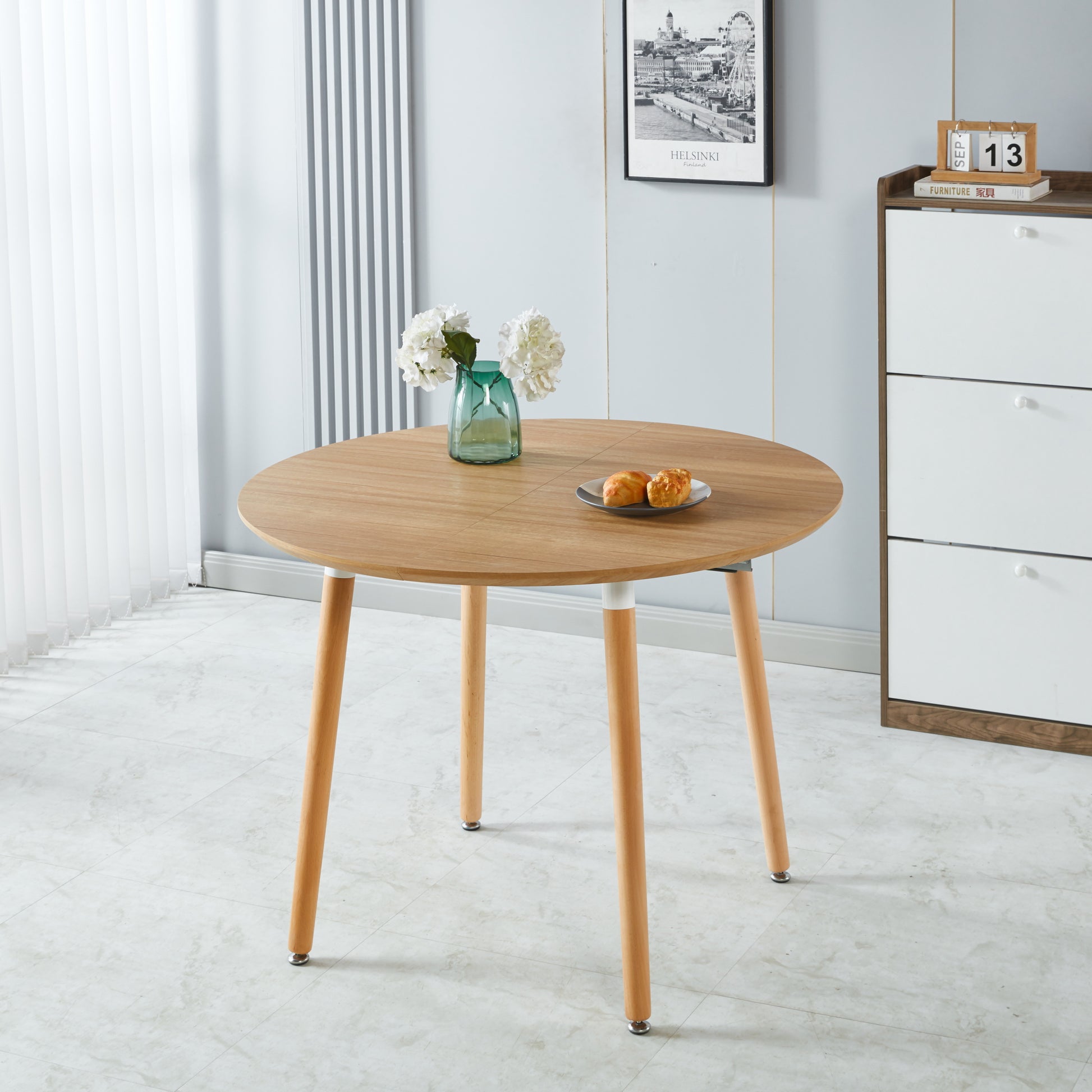 Round oak wood dining table with light wood legs featuring a vase of white flowers and pastries