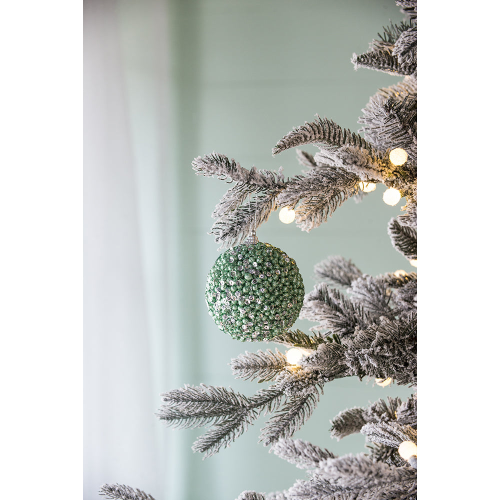 Glittering green Christmas ornament with sequin details hanging on a flocked pine branch