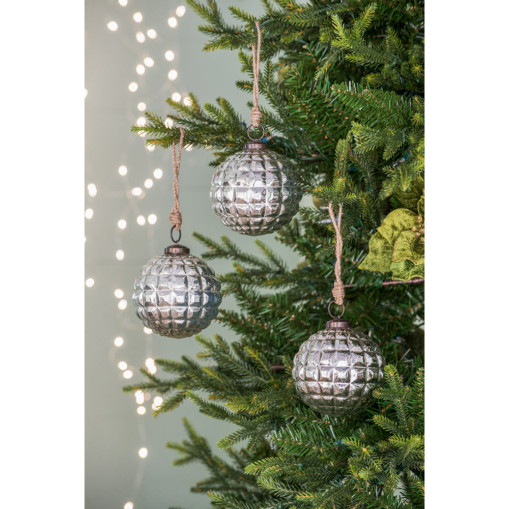 Three textured silver glass Christmas ornaments hang on a pine tree