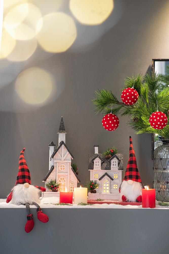 Christmas gnome with red and black buffalo plaid hat and fuzzy beard sits beside holiday village houses