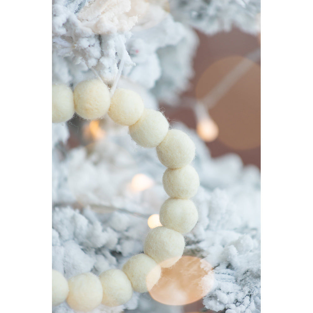 Cream felt ball garland draped on a frosted Christmas tree with warm bokeh lights