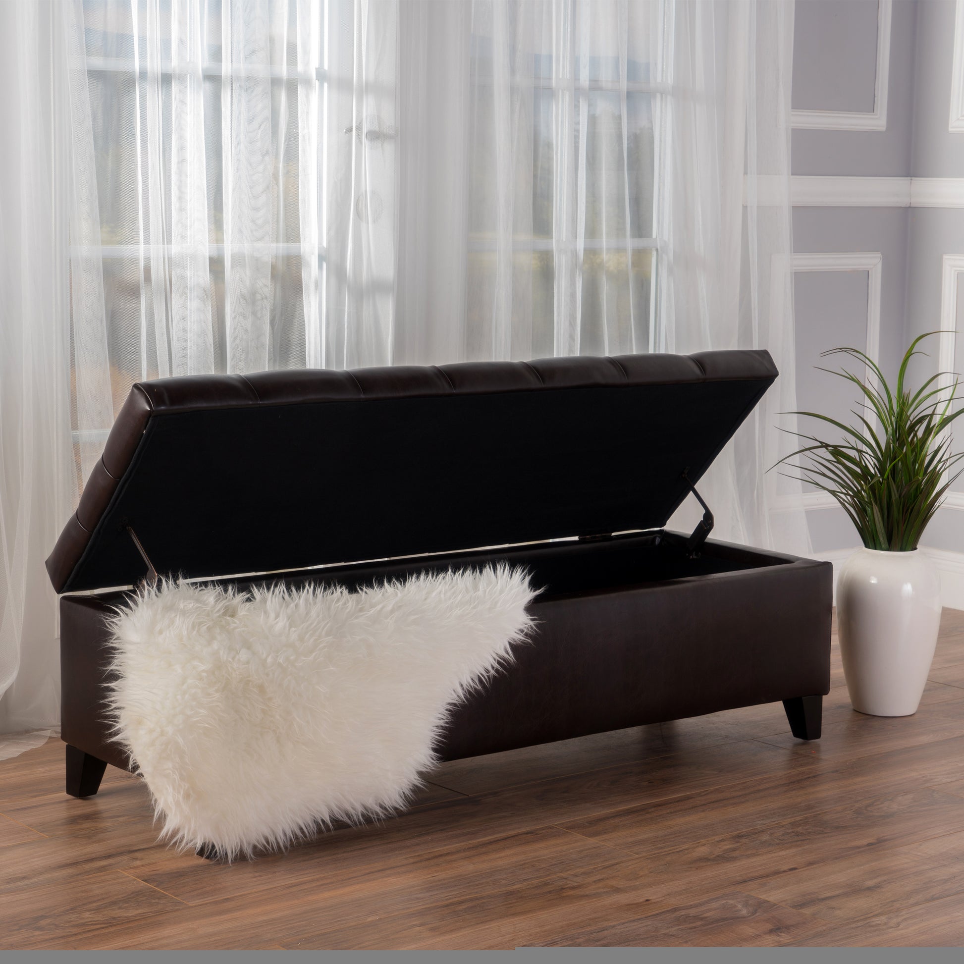 Dark brown faux leather storage ottoman with tufted lid open revealing black interior and white fluffy throw