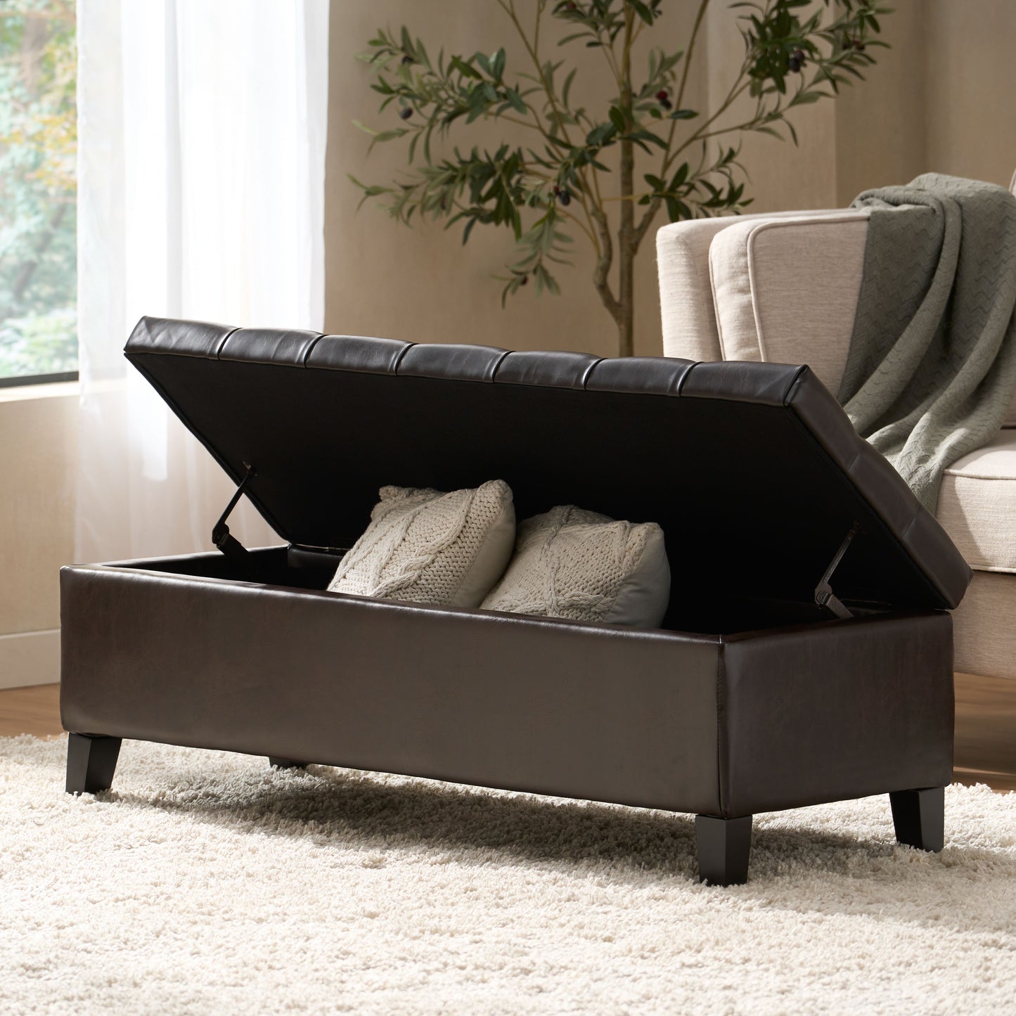 Dark brown faux leather storage ottoman with tufted lid and two cream knit pillows inside