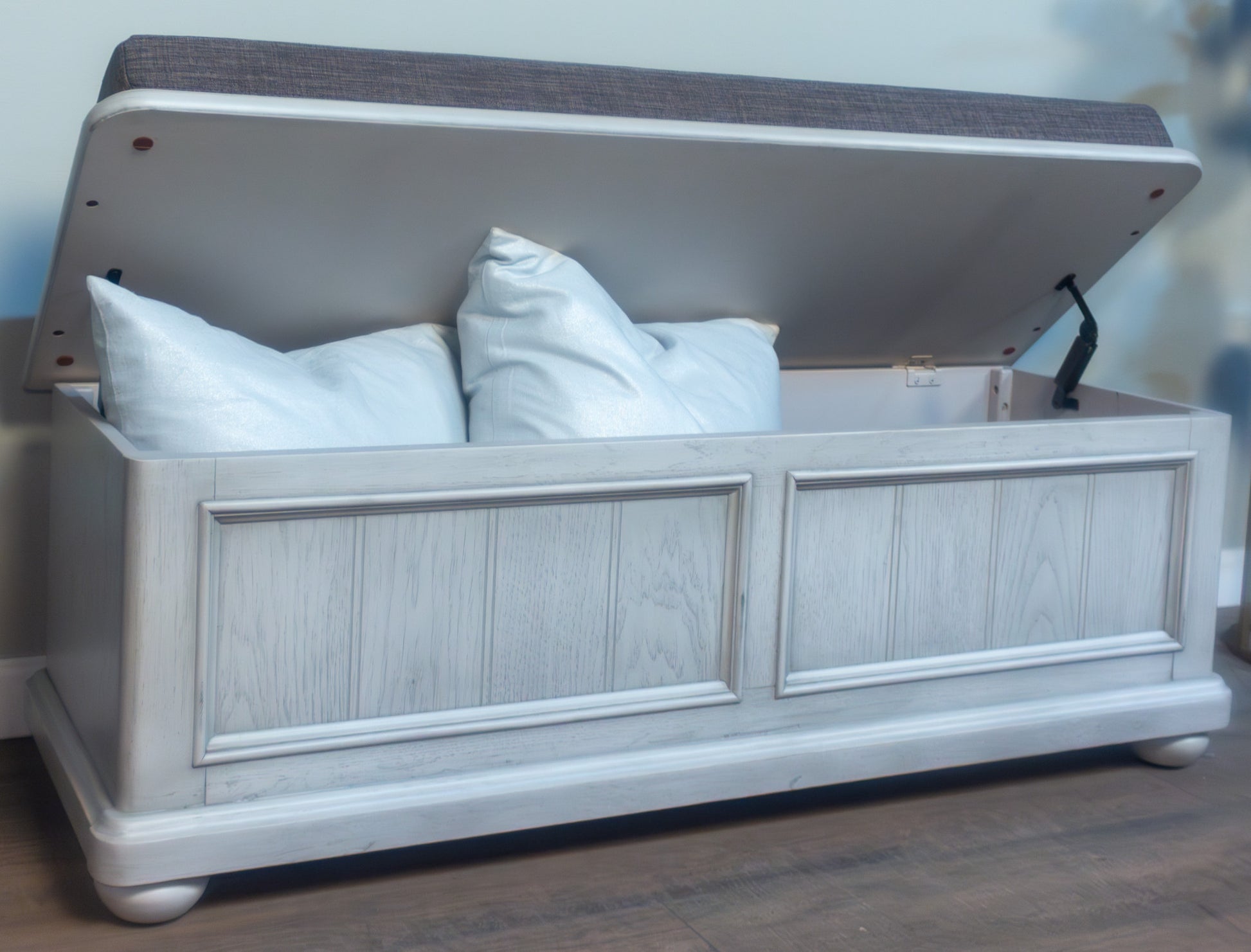 Light grey wooden storage ottoman with hinged upholstered lid and light blue pillows inside